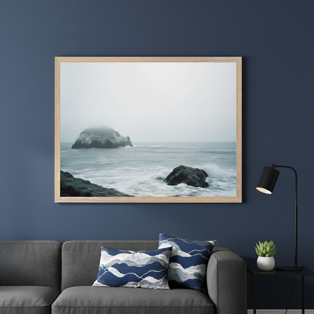 Modern living room with a dark gray sofa. A large light wood framed print of a foggy ocean scene with rock formations and crashing waves hangs above the sofa. Blue patterned pillows are on the sofa, and a black lamp is on a side table with a succulent.