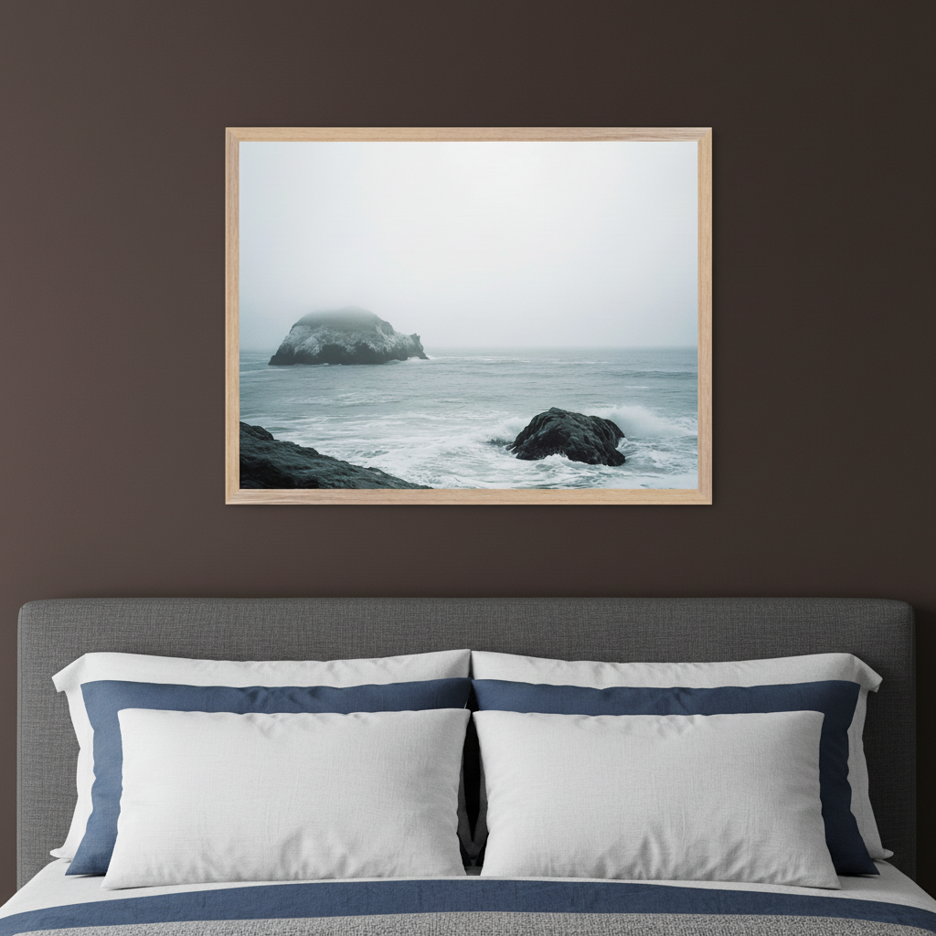 Modern bedroom with a gray upholstered bed and white and blue pillows. A light wood-framed photograph of a foggy, rocky ocean scene with waves crashing on dark rocks hangs centered on the dark brown wall above the bed.