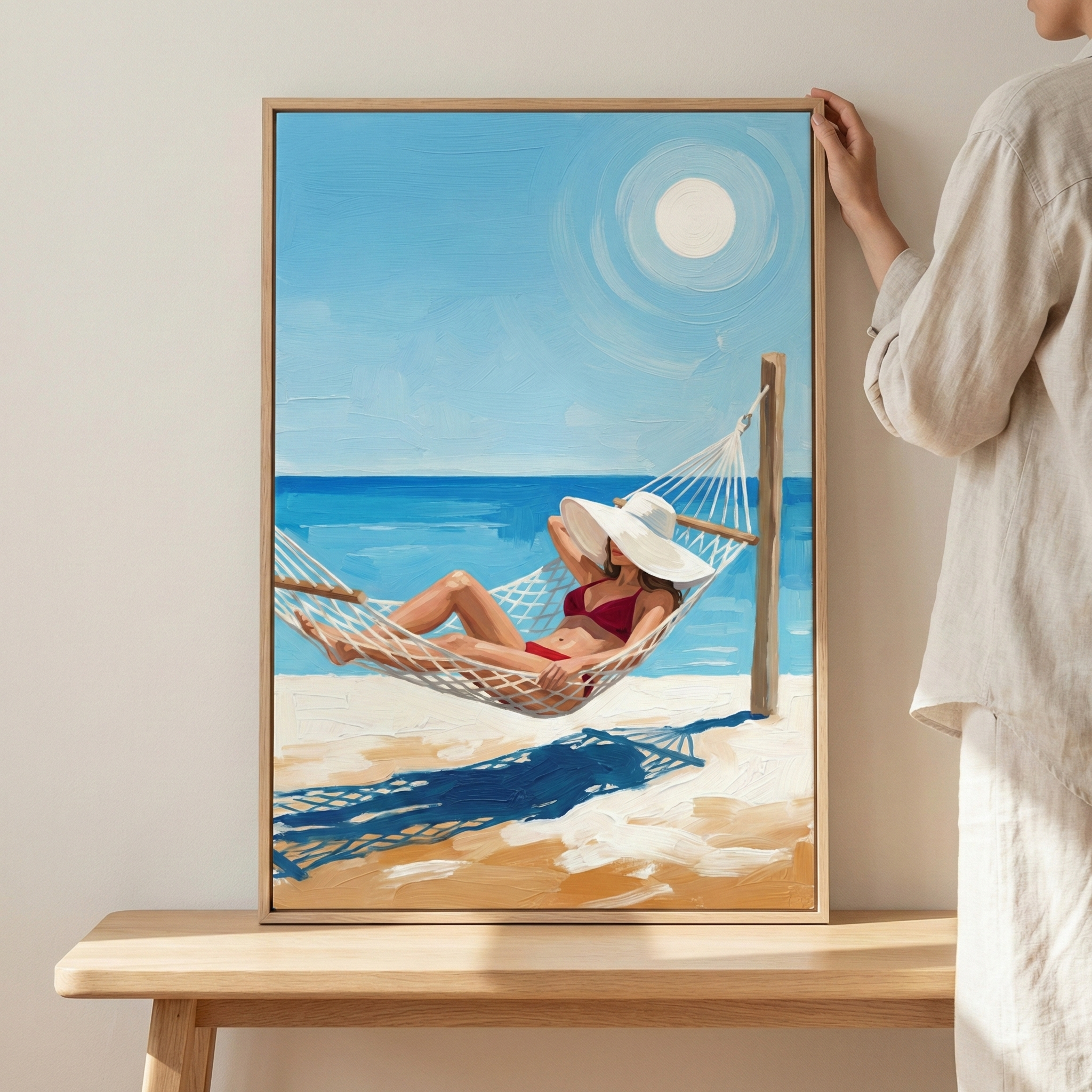 A person stands beside a large, vertically oriented framed artwork resting on a light-colored wooden table. The artwork features a woman in a red bikini and a wide-brimmed white hat, relaxing in a white mesh hammock on a bright beach. The composition is created with thick, expressive brushstrokes, depicting a vibrant blue ocean and a clear sky with a large white sun. The hammock casts a prominent dark blue patterned shadow across the sandy foreground. The piece is encased in a simple light wood frame.
