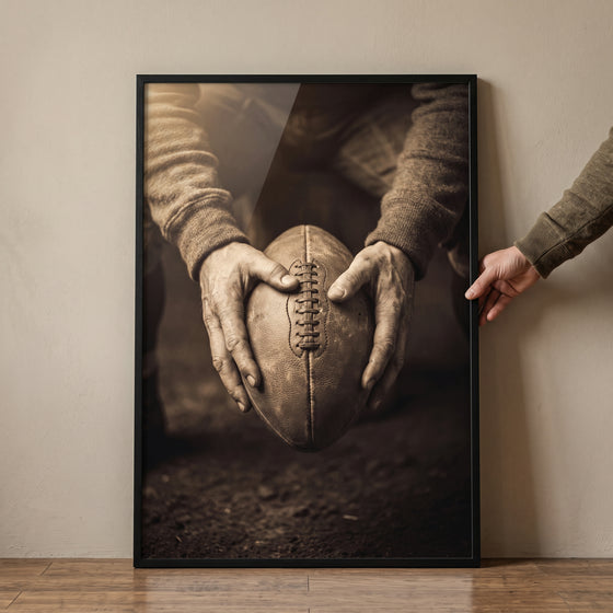 Framed sepia hands holding football sports wall art leaning on a wall, held by a hand.