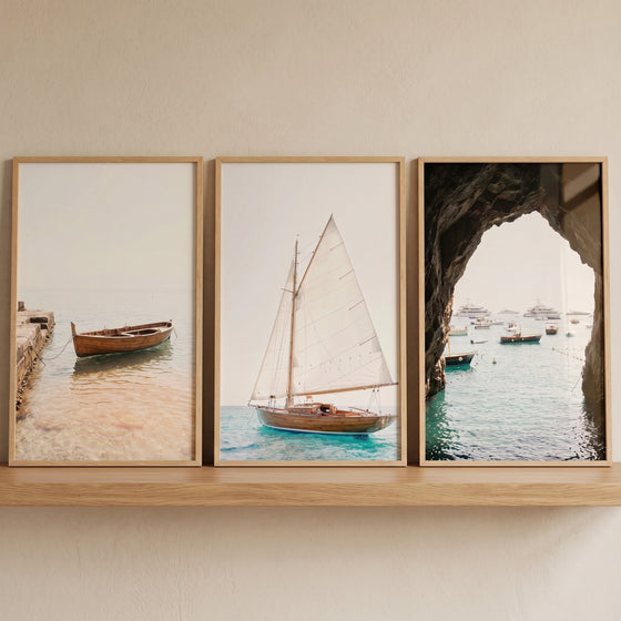 Serene blue water boat artwork set of 3 framed on a shelf, showing a rowboat, a sailboat, and a view from a sea cave.