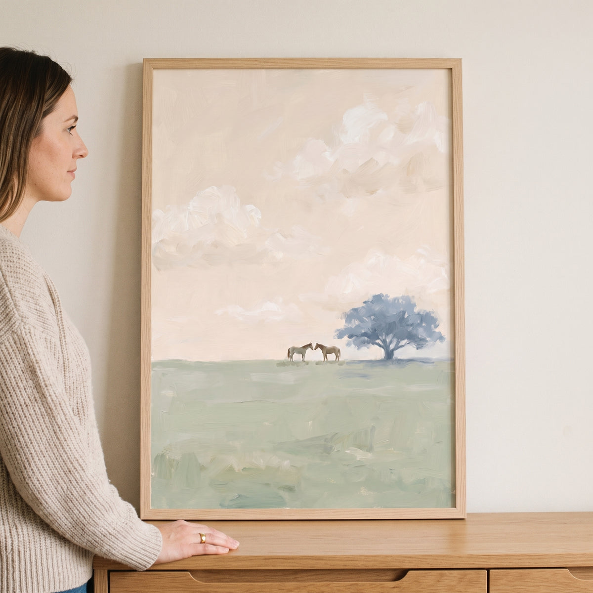 Woman stands by serene field tree horses wall art showing a blue tree and two horses in a soft, minimalist landscape.