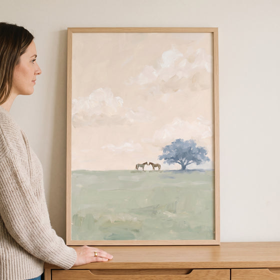 Woman stands by serene field tree horses wall art showing a blue tree and two horses in a soft, minimalist landscape.