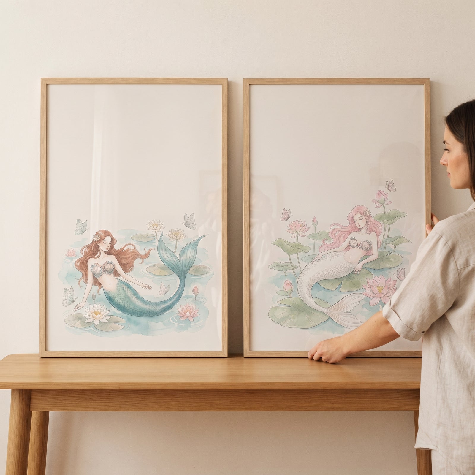 A woman adjusts a serene floating lotus mermaid wall art set of 2 in light wood frames resting on a wooden table.