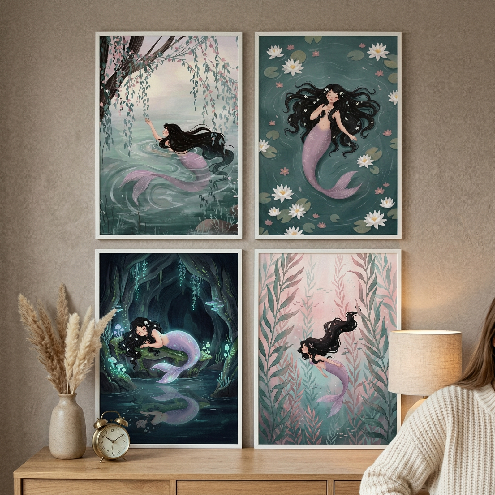 Four framed mermaid illustrations displayed on a wooden dresser showing soft underwater scenes with flowing dark hair lily pads caves and sea plants in muted pastel tones. A vase with dried grasses and a small clock sit beside the prints while part of a person in a cozy sweater is visible creating a warm styled home decor setting.