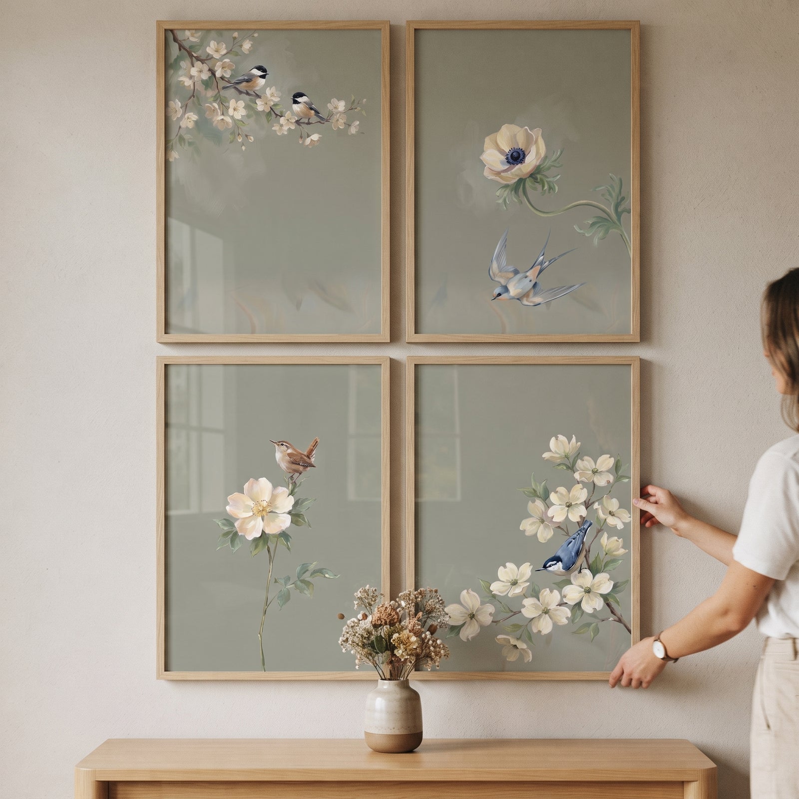 A woman adjusts a serene muted garden bird wall art set of 4 in light wood frames on a wall above a flower vase.