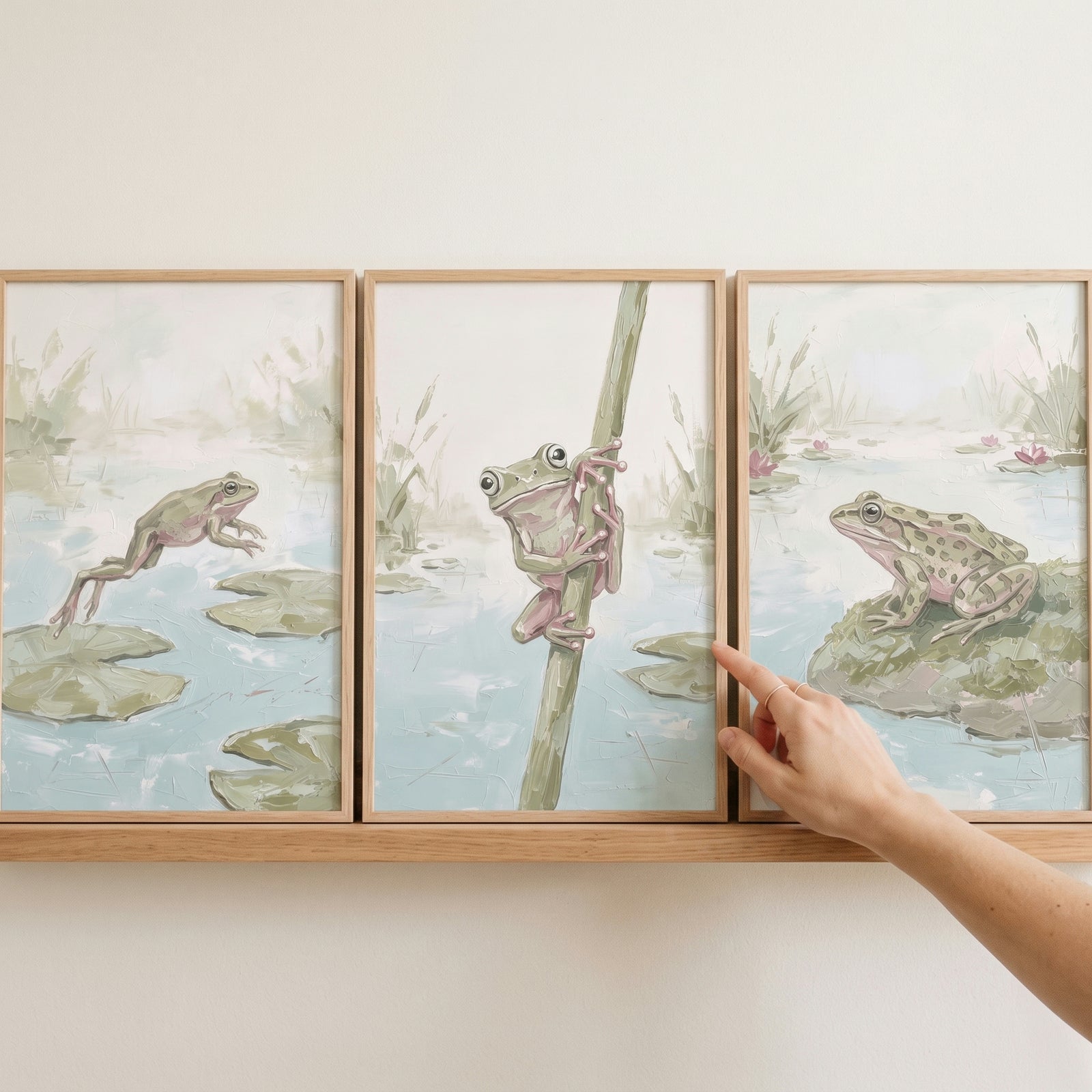 Serene sage green frog wall art set of 3 framed prints sits on a wooden shelf as a person adjusts a frame.