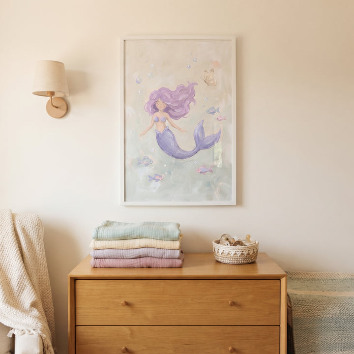 Serene swimming pastel mermaid wall art hangs above a wooden dresser with folded blankets in a soft-toned nursery.