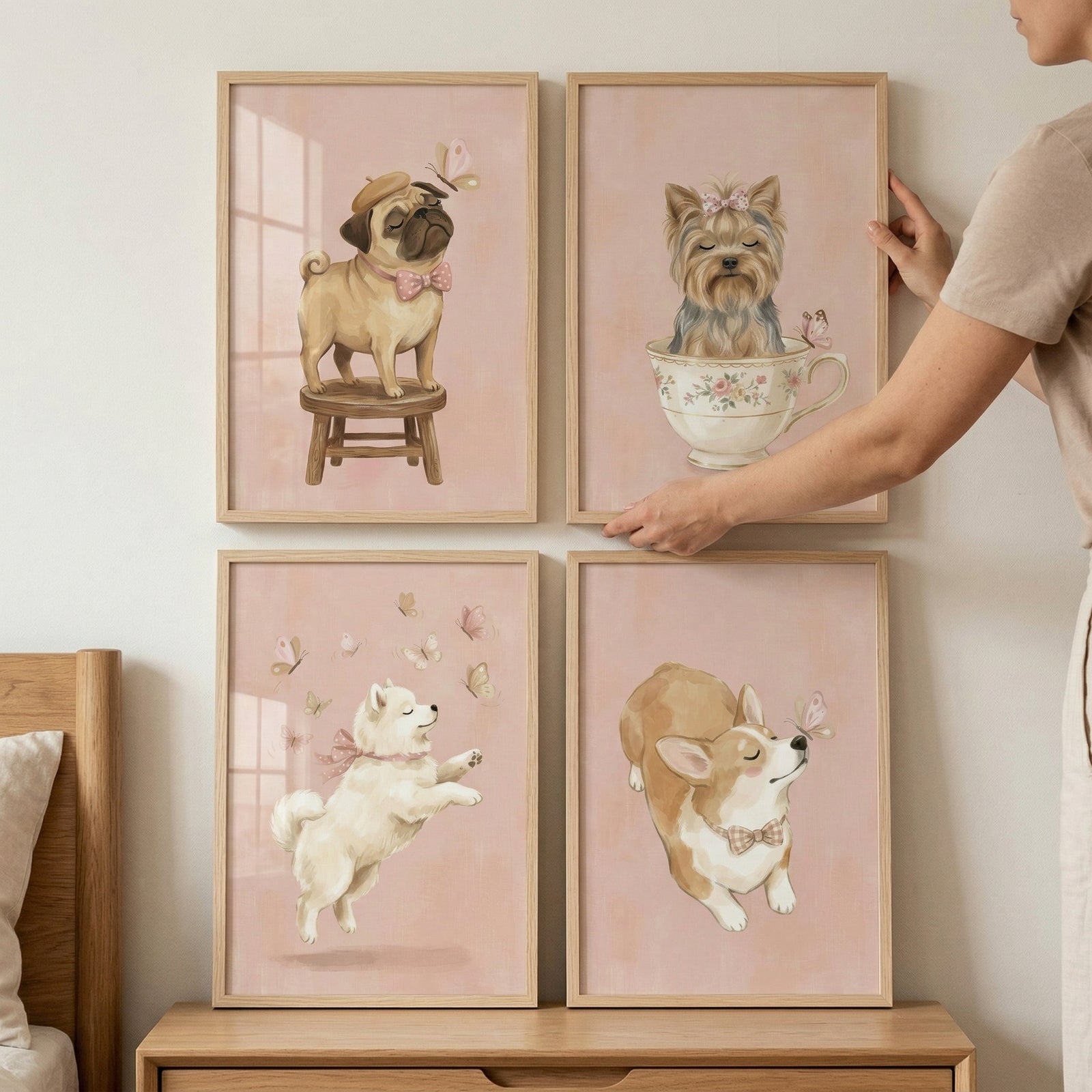 A collection of four framed illustrations of whimsical dogs is displayed on a light-colored wall in a two-by-two grid. Each piece of art features a soft pink background and is encased in a simple light wood frame.

In the top-left frame, a pug wearing a small beret and a pink bow tie stands on a three-legged wooden stool. The top-right frame, which a person's hand is shown adjusting, depicts a small, long-haired dog nestled inside an ornate floral teacup.
