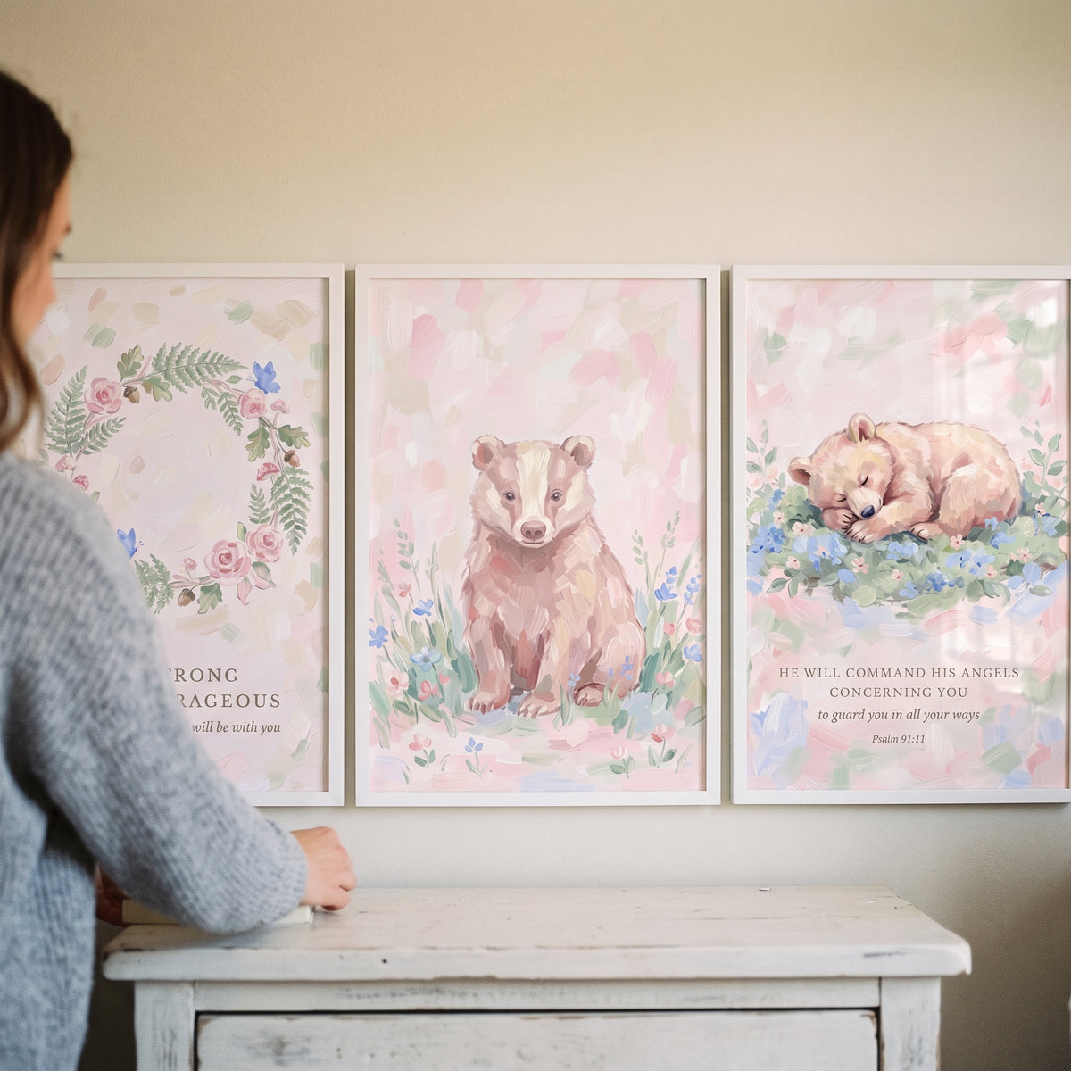 Woman stands by a shabby chic pink woodland artwork set of 3 featuring a wreath, badger, and bear above a white dresser.