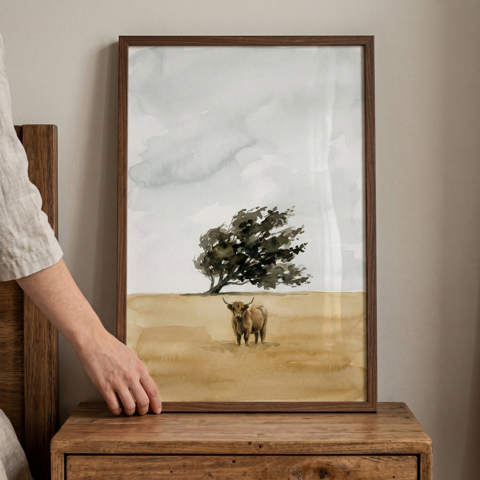 A hand holds a framed shaggy brown highland cow wall art featuring a lone cow and tree in a muted watercolor landscape.
