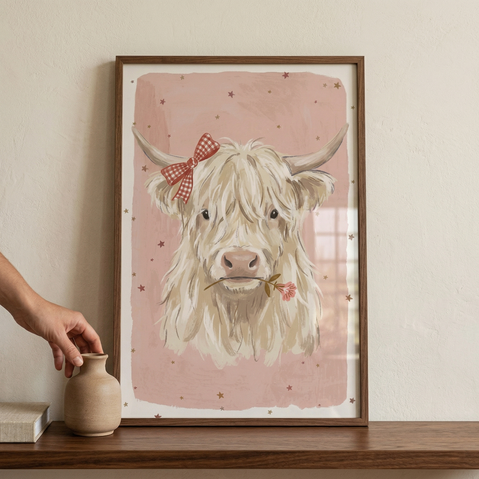 A vertical, framed illustration features a portrait of a long-haired, cream-colored cow. The cow is adorned with a red and white checkered bow on its horn and holds a delicate pink flower in its mouth. The background of the artwork is a textured, dusty-pink color with small, scattered star shapes. The piece is set in a dark wood frame and rests on a dark wooden surface against a plain, off-white wall. To the left, a hand reaches toward a small, neutral-toned ceramic vase.