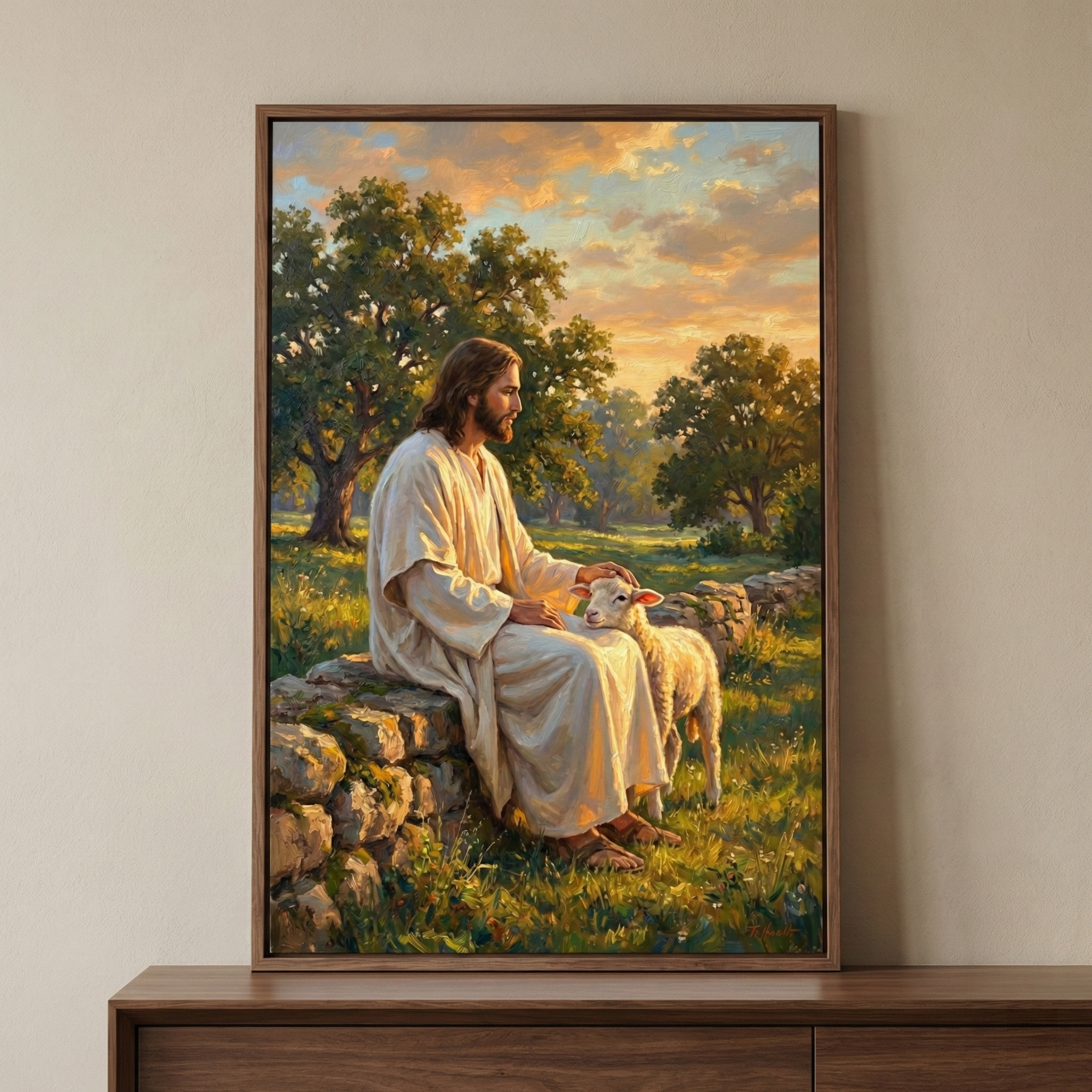 This framed artwork depicts Jesus Christ sitting peacefully on a stone wall in a rolling green field. He is dressed in simple, off-white robes and has long brown hair and a beard. With a kind expression, he gently pets a small white lamb standing next to him. The background features large trees under a vast sky filled with warm, golden clouds at sunset. The soft, radiant light creates a tranquil and reverent atmosphere throughout the entire scene.