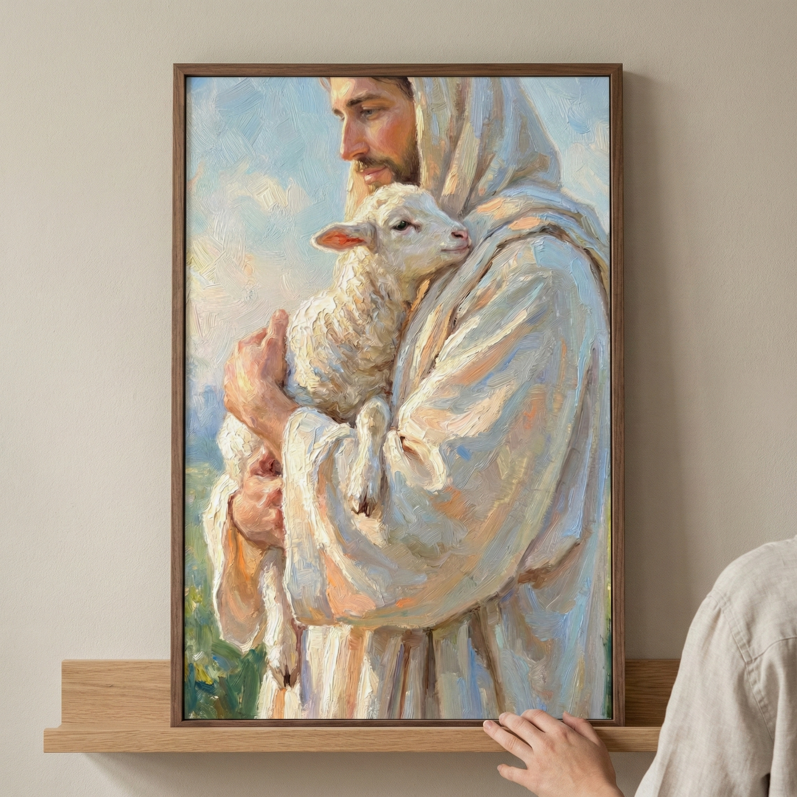 A framed vertical piece of art on a wooden shelf depicts Jesus Christ gently holding a small lamb close to his chest. Jesus is shown in profile with a beard and a calm expression, wearing a flowing white robe and head covering detailed with thick, textured strokes of light blue and yellow. The small white lamb rests its head against him. The background consists of soft blue and white hues, and a hand is visible at the bottom right corner touching the shelf.