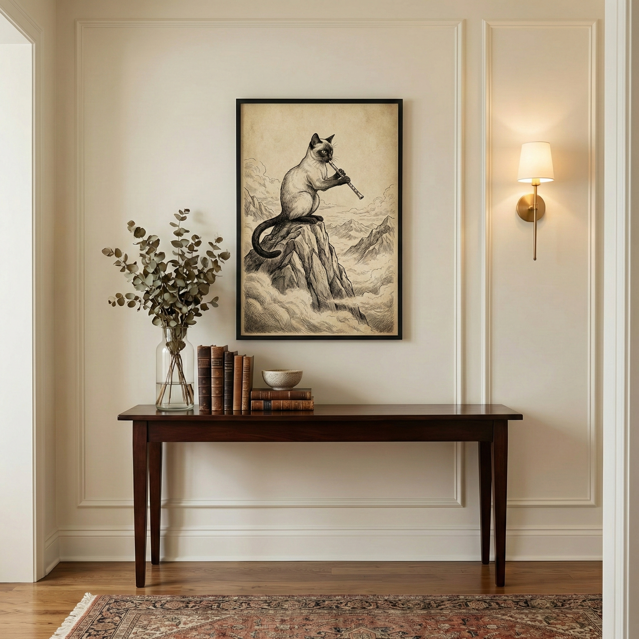 An interior scene featuring a cream-colored wall with traditional molding. At the center is a framed vintage-style illustration of a Siamese cat perched on a jagged mountain peak, playing a flute. Below the art, a dark wood console table holds a glass vase with eucalyptus, several old books, and a small bowl. A gold wall sconce with a white shade is mounted to the right, emitting a warm light. An ornate rug lies on the wood floor at the bottom of the frame.