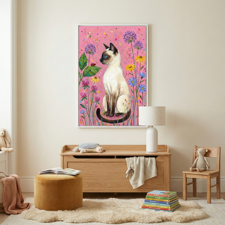 A framed piece of artwork hangs on a light-colored wall. The illustration features a cream-colored cat with dark points sitting among colorful wildflowers on a bright pink background. Below, a wooden storage chest holds a white lamp and a plush toy. In the foreground, a yellow velvet ottoman with an open book sits on a shaggy rug next to a stack of colorful books. A small wooden chair with a plush toy is on the right. The room is softly lit and cozy.