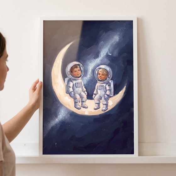 A hand holds a framed sitting moon baby astronauts wall art featuring two children in spacesuits on a crescent moon.