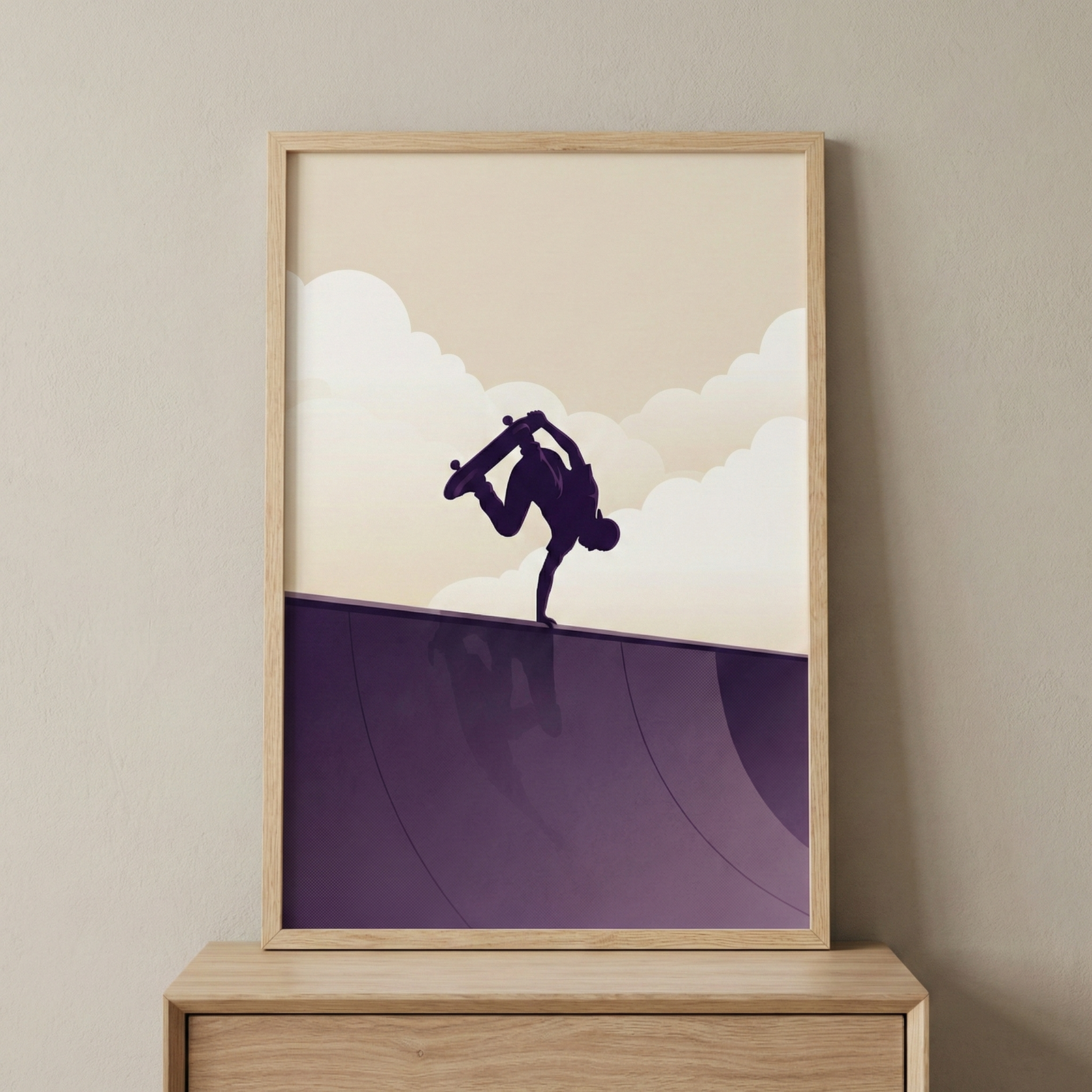 A framed minimalist illustration depicts the dark silhouette of a skateboarder performing a handplant trick on the edge of a curved purple ramp. Above, stylized clouds float in a cream sky. A subtle reflection of the figure is visible on the ramp's surface. The entire scene uses a simple purple and off-white color palette. This artwork is presented in a light-toned wooden frame, resting on a matching wooden surface against a neutral, flat-colored wall.