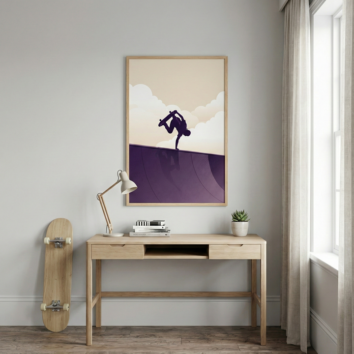 A vertical framed art print hangs on a light grey wall above a minimalist wooden desk. The illustration in the frame depicts a silhouette of a person performing a handplant on a skateboard atop a purple curved ramp, set against a cream-colored sky with stylized white clouds. On the desk, there is a small white lamp with a curved neck, a stack of books, and a tiny green plant in a white pot. To the left of the desk, a natural wood skateboard leans against the wall.