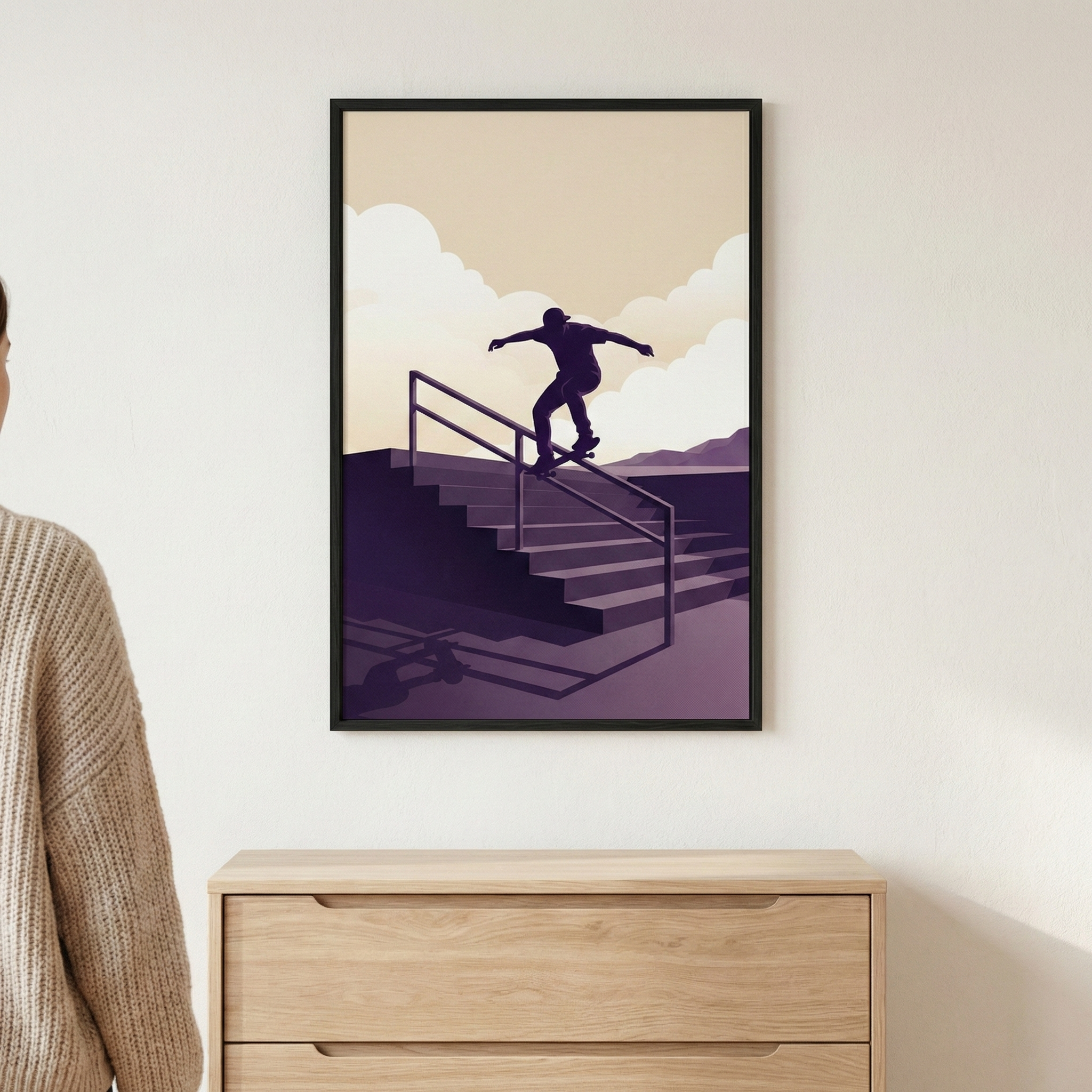 A minimalist framed graphic illustration hangs on a light-colored wall, positioned above a wooden chest of drawers. The artwork features a dark silhouette of a skateboarder performing a grind on a handrail next to a flight of stairs. The scene is rendered in a limited color palette of deep purple, cream, and tan. Stylized, billowy clouds fill the sky behind the figure, with distant purple hills on the horizon.