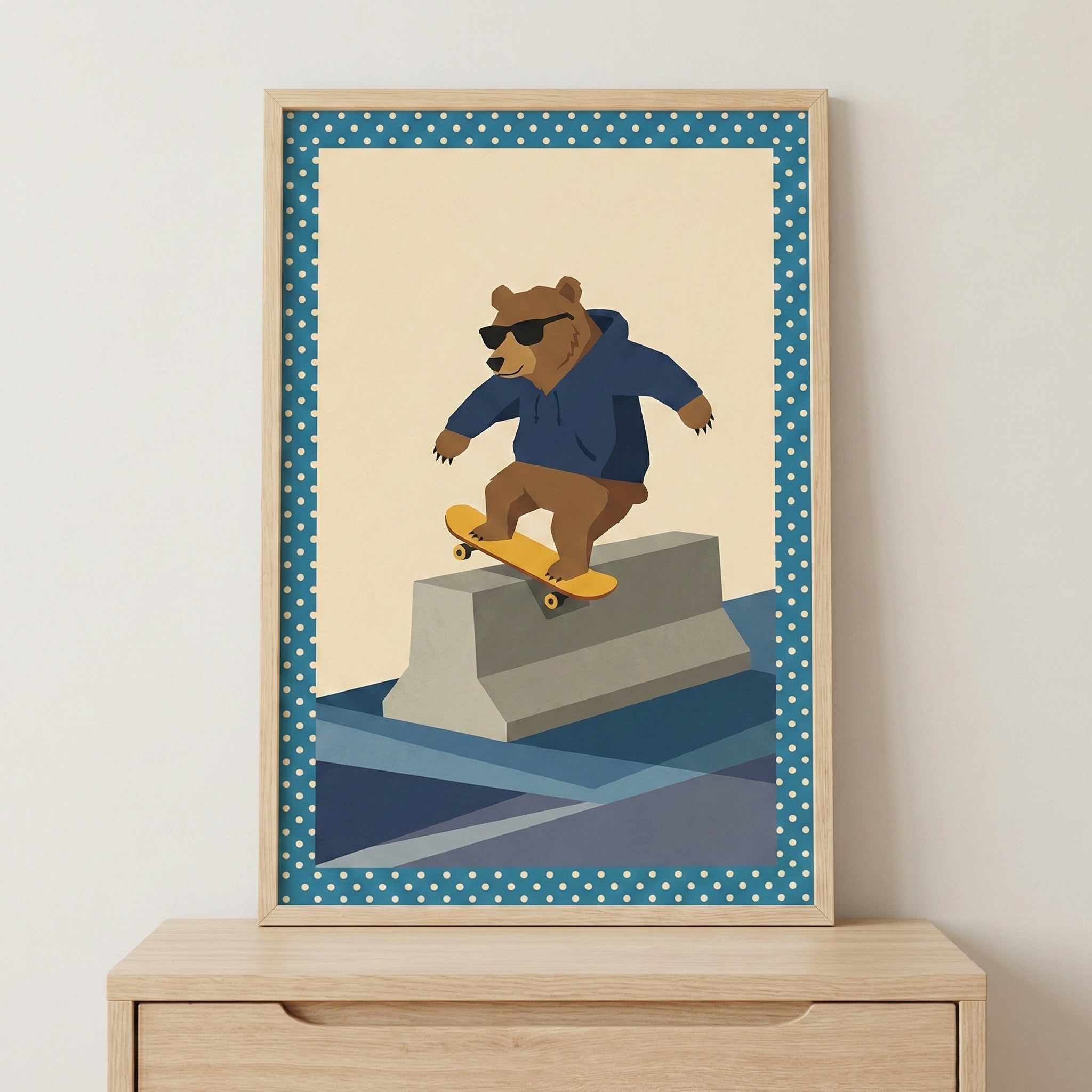 A framed graphic illustration of a brown bear wearing a dark blue hooded sweatshirt and black sunglasses. The bear is skateboarding on a yellow skateboard, sliding across the top of a gray concrete barrier. The illustration features a minimalist style with a cream background and blue geometric shapes for the ground. A teal border with white polka dots surrounds the image, which is set in a light wooden frame and displayed on top of a light-colored wood cabinet.