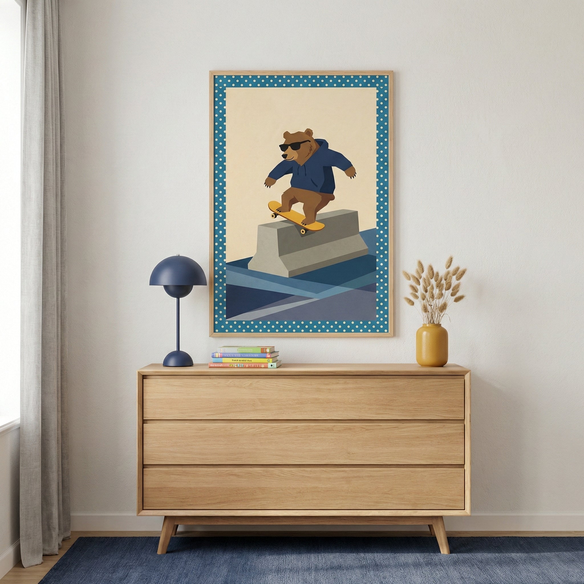 A framed illustration of a brown bear wearing a blue hoodie and sunglasses while skateboarding on a concrete barrier is mounted on a light-colored wall above a wooden three-drawer dresser. The artwork has a blue border with white polka dots. On the dresser, there is a small navy blue lamp, a short stack of books, and a mustard yellow vase filled with dried botanicals. A dark blue rug is on the floor, and a light-colored curtain hangs to the left.