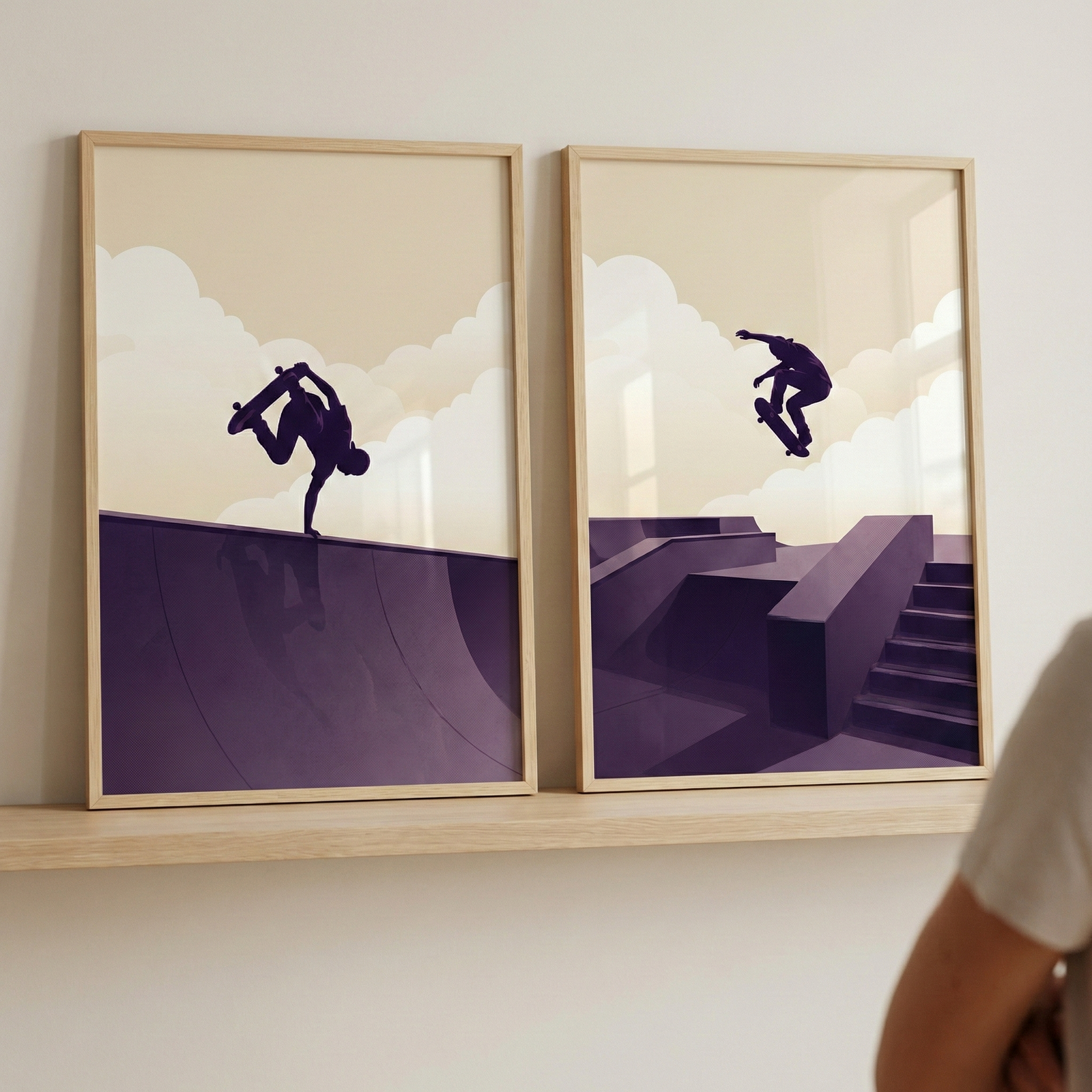 Two framed art prints sit side-by-side on a light wood shelf against an off-white wall. The prints feature a minimalist style with a palette of deep purple, cream, and tan. The left print shows a dark silhouette of a person doing a handplant on a skateboard atop a curved purple ramp. The right print displays a silhouette of a person mid-air on a skateboard jumping over purple skatepark ledges and stairs. Both have backgrounds of cream-colored clouds against a tan sky.