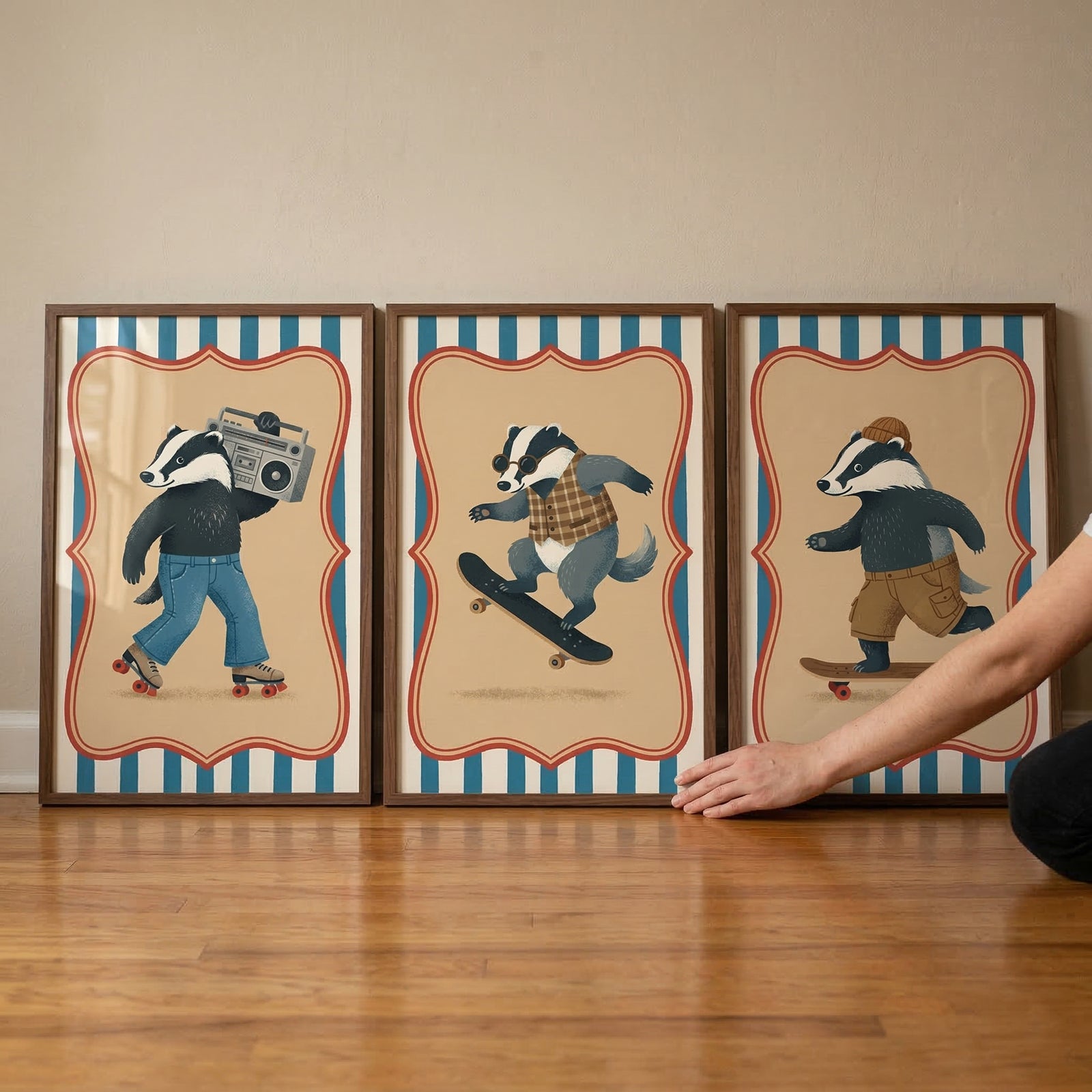 Three framed illustrations of badgers in various urban settings are lined up on a light-colored wooden floor. Each piece shows a badger character within a tan-bordered frame against blue and white vertical stripes. From left to right: a badger in blue pants on roller skates carries a portable music player; a badger in a plaid vest and sunglasses performs a skateboard trick; and a badger wearing a beanie and shorts rides a skateboard. A hand is seen adjusting the frame on the right.