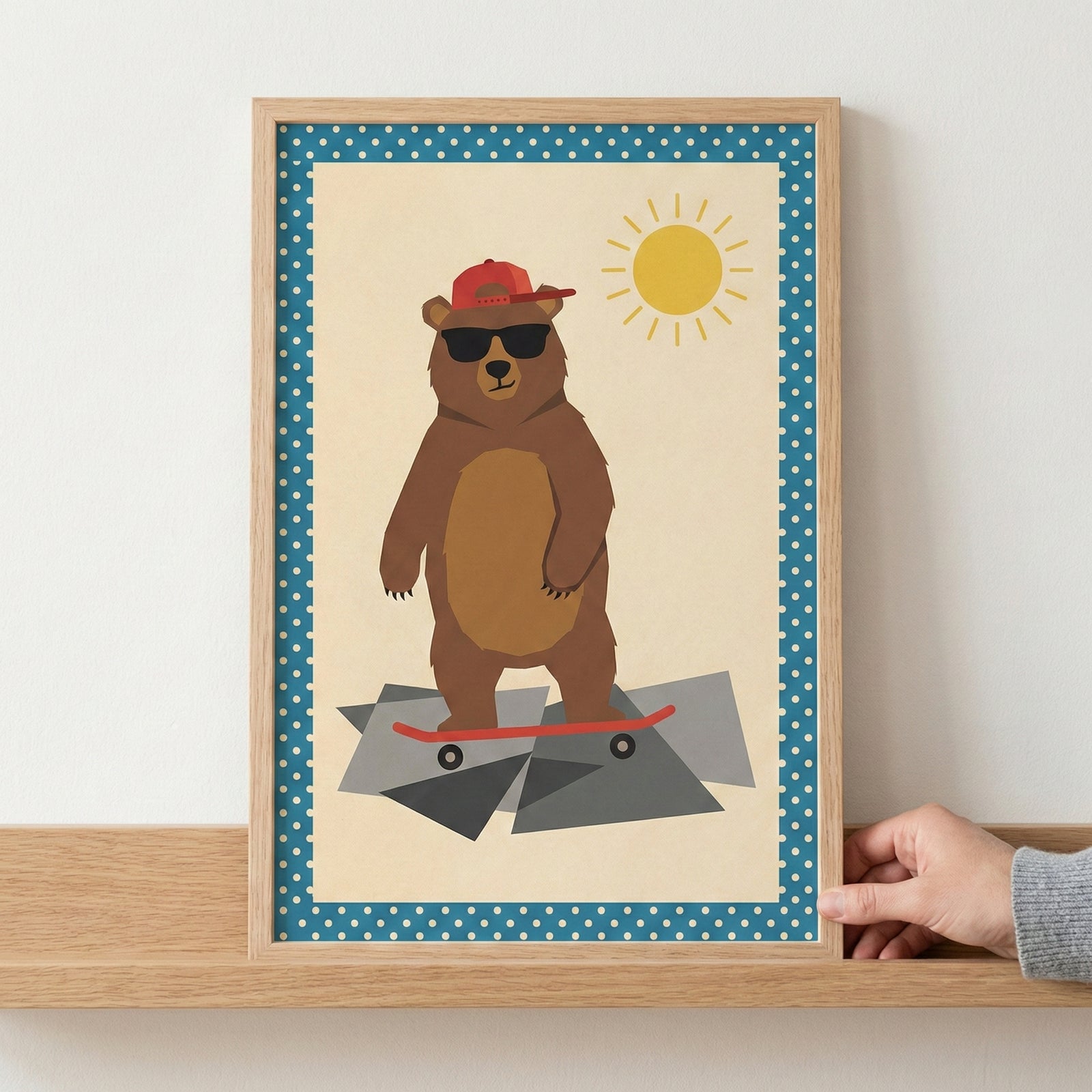 A framed illustration of a brown bear wearing black sunglasses and a red baseball cap, standing on a red skateboard. The bear is depicted on a jagged gray surface against a pale background with a simple yellow sun in the upper corner. The artwork features a blue border with white polka dots and is shown in a light wood frame resting on a wooden shelf.