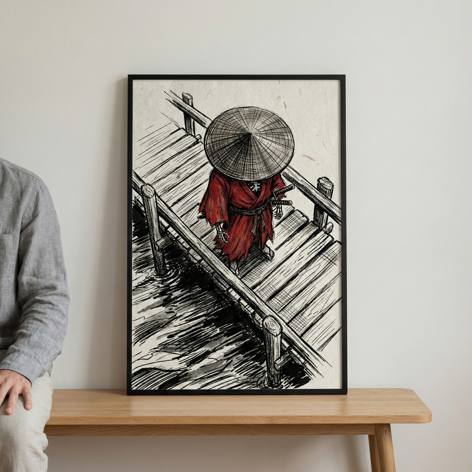 A framed ink illustration rests on a light wood table against a plain wall. The artwork depicts a figure in a bright red robe and a wide conical straw hat, viewed from an elevated perspective. The figure stands on a wooden pier above water rendered with bold black strokes. Two swords are visible at the figure's side. To the left, a person in a gray long-sleeved shirt is partially visible, sitting on the edge of the table.