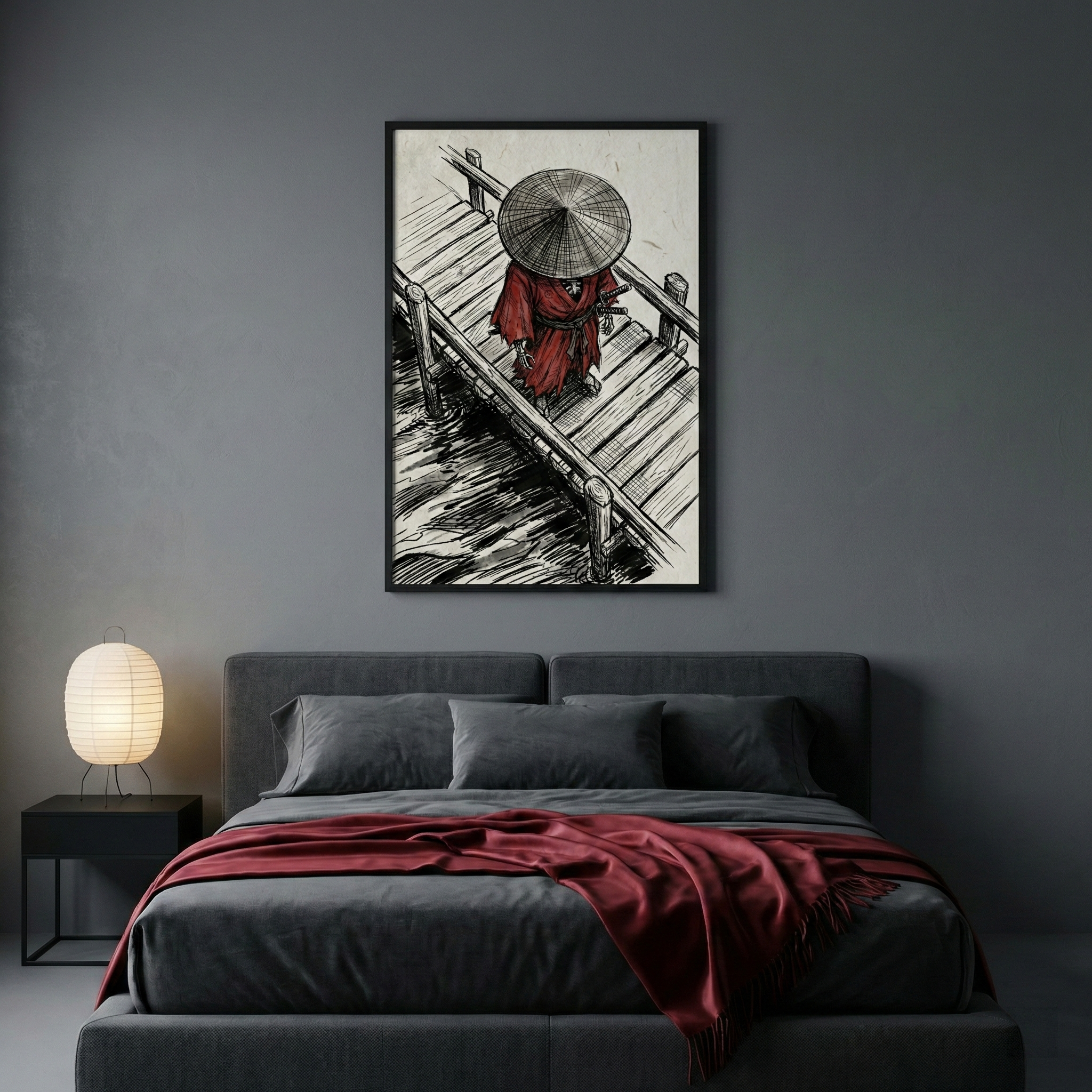 A modern, minimalist bedroom features a dark grey wall and a charcoal-upholstered bed. On the bed, a deep red throw blanket adds a pop of color alongside matching grey pillows. To the left, a small black bedside table holds a spherical paper lantern lamp. Centered above the headboard is a large framed piece of art showing a figure in a red robe and a wide, conical straw hat standing on a wooden dock over water, rendered in a high-contrast ink drawing style.
