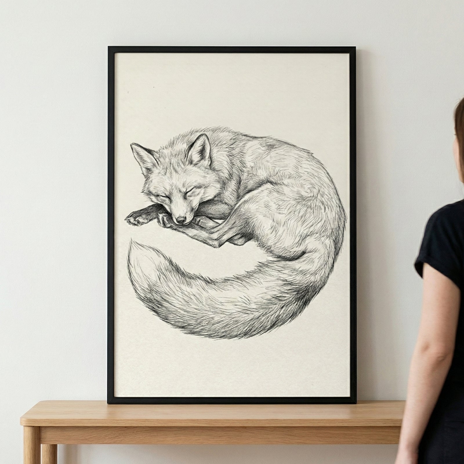 A detailed black and white sketch of a fox curled up in a sleeping position. The artwork is presented in a simple black frame and rests on a light-colored wooden surface against a plain wall. A person's shoulder and arm are partially visible to the right.