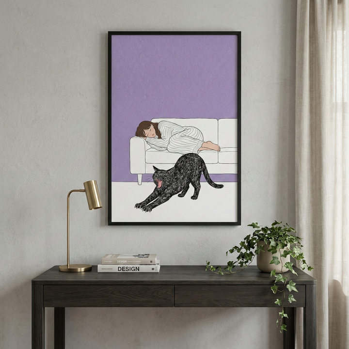 A framed illustration hangs on a light gray wall above a dark wood desk. The artwork depicts a woman with brown hair sleeping on a white sofa against a solid lavender background. In the foreground of the art, a black cat stretches its body across a white floor. On the desk below, there is a modern gold-toned lamp, a small stack of books with the word "DESIGN" on the spine, and a potted plant with green vines trailing down. To the right, soft white curtains hang near a window.
