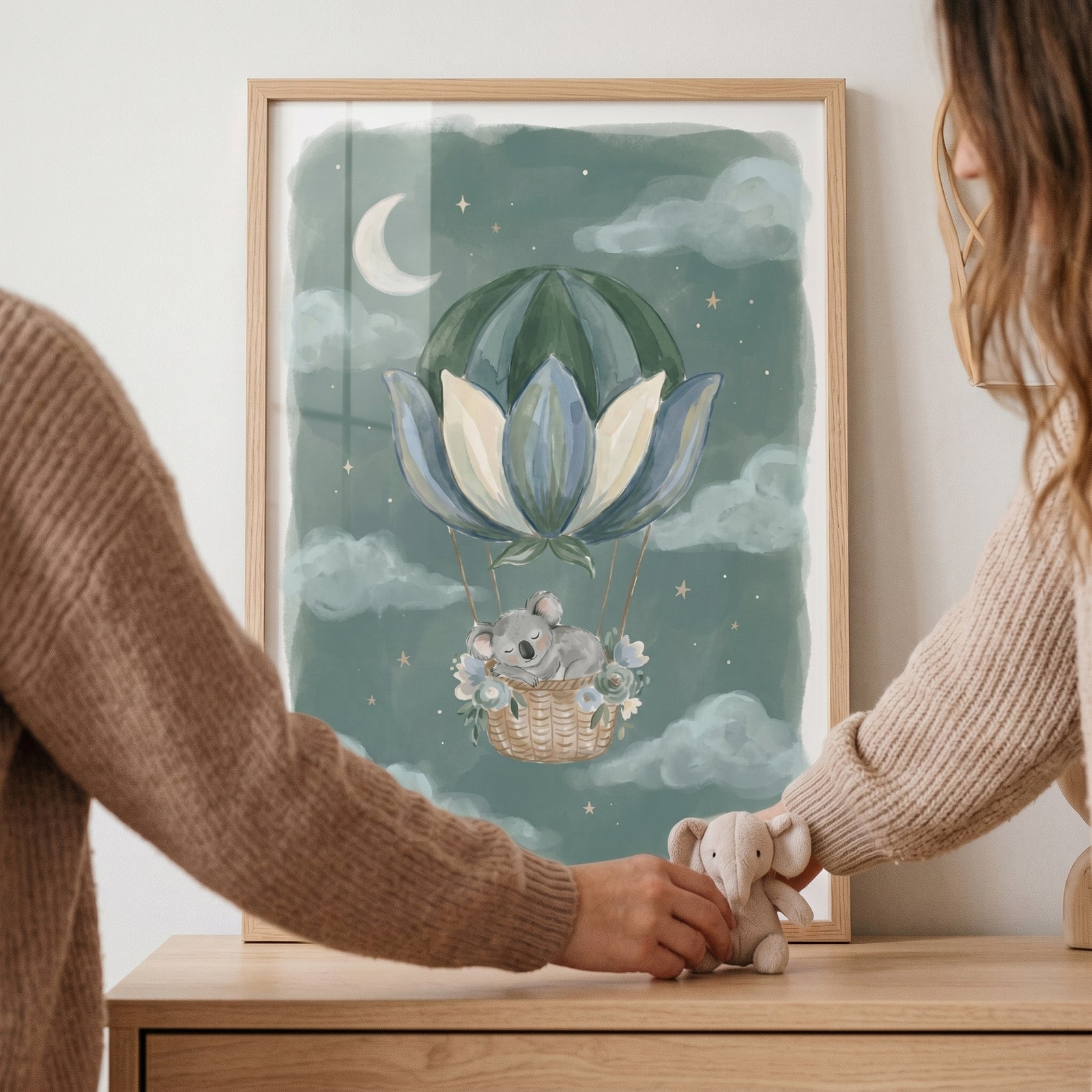 Framed sleeping lotus balloon koala wall art held by two people next to a small stuffed elephant on a table.