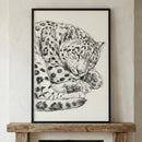 A black and white sketch of a snow leopard curled up and sleeping. The detailed illustration captures the feline's thick, spotted coat and large paws tucked under its head. The artwork is set in a simple black frame and rests on a rustic wooden mantelpiece against a light-colored, textured wall.