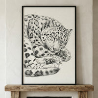 A black and white sketch of a snow leopard curled up and sleeping. The detailed illustration captures the feline's thick, spotted coat and large paws tucked under its head. The artwork is set in a simple black frame and rests on a rustic wooden mantelpiece against a light-colored, textured wall.