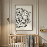 A framed black and white sketch of a sleeping leopard hangs on a paneled wall above a light-colored wooden crib in a neutral-toned nursery. A plush llama toy and a grey knitted blanket rest inside the crib. To the left, a tall gold floor lamp with a slim head illuminates the corner. On the right, a small wooden wall shelf holds several children's books.