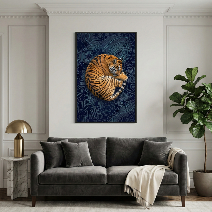 A modern living room features a striking piece of wall art as its centerpiece. The artwork depicts a realistic tiger curled into a circular pose against a dark blue background filled with luminous, swirling contour lines. Below, a dark grey velvet sofa is adorned with plush pillows and a cream-colored knit throw. A marble side table with a sleek gold lamp sits to the left, while a large, leafy green plant in a pot stands to the right. The white walls are accented with elegant molding.