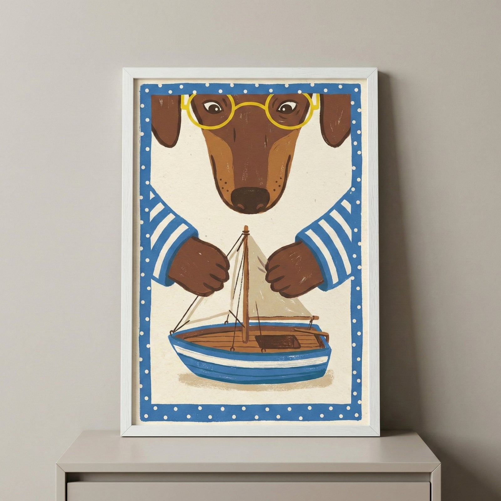 A framed illustration features a brown dachshund with yellow glasses and a blue-and-white striped shirt. The dog appears to be carefully holding or building a small wooden sailboat with a white sail. The artwork is framed by a blue border with white polka dots and is displayed in a white frame on a light-colored surface against a neutral wall.
