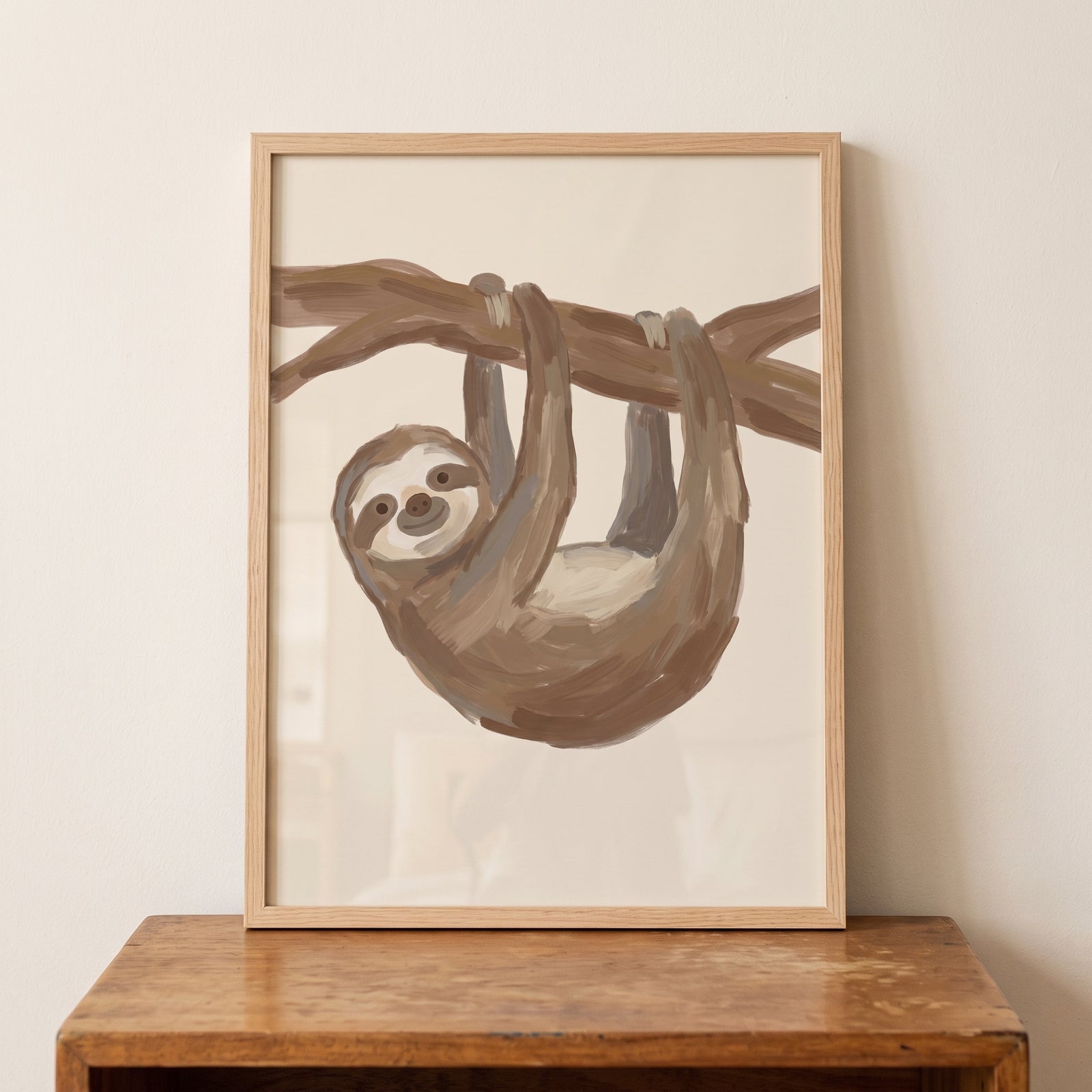 Framed smiling brown baby sloth wall art hanging on a branch, resting on a wooden table against a beige wall.