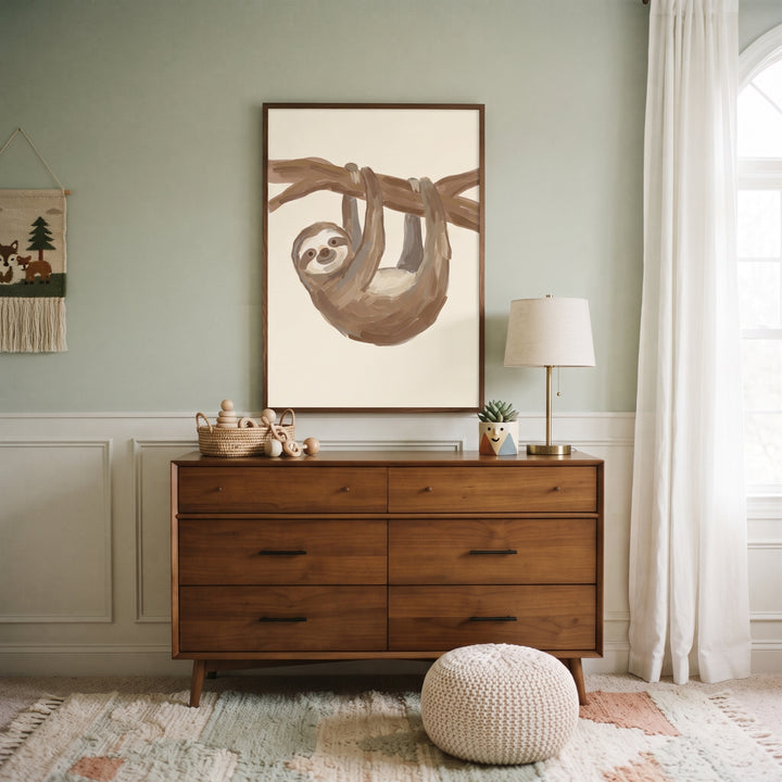 Smiling brown baby sloth wall art hangs above a wooden dresser in a modern nursery with a textured rug and knit poof.