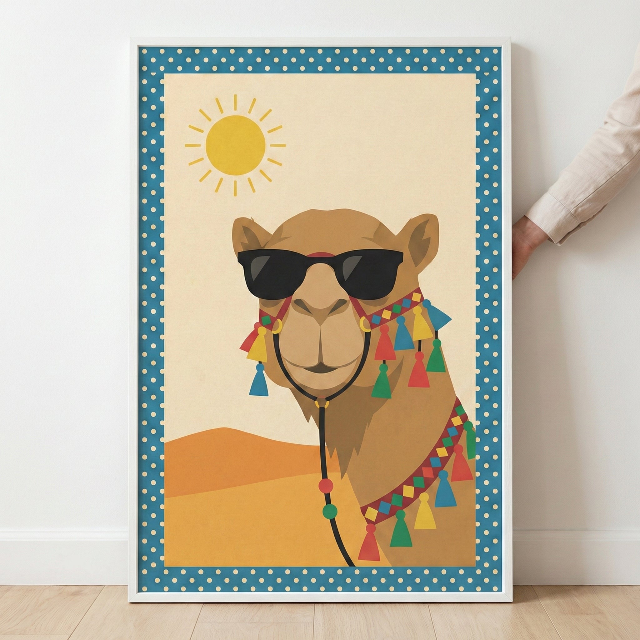 A framed vertical art print featuring a stylized illustration of a camel wearing black sunglasses and a colorful, tasseled harness. The background depicts a simple desert scene with rolling sand dunes under a bright yellow sun. The central artwork is bordered by a blue frame with white polka dots. The white-framed print leans against a white wall on a light wood floor, with a person's hand visible holding the side of the frame.