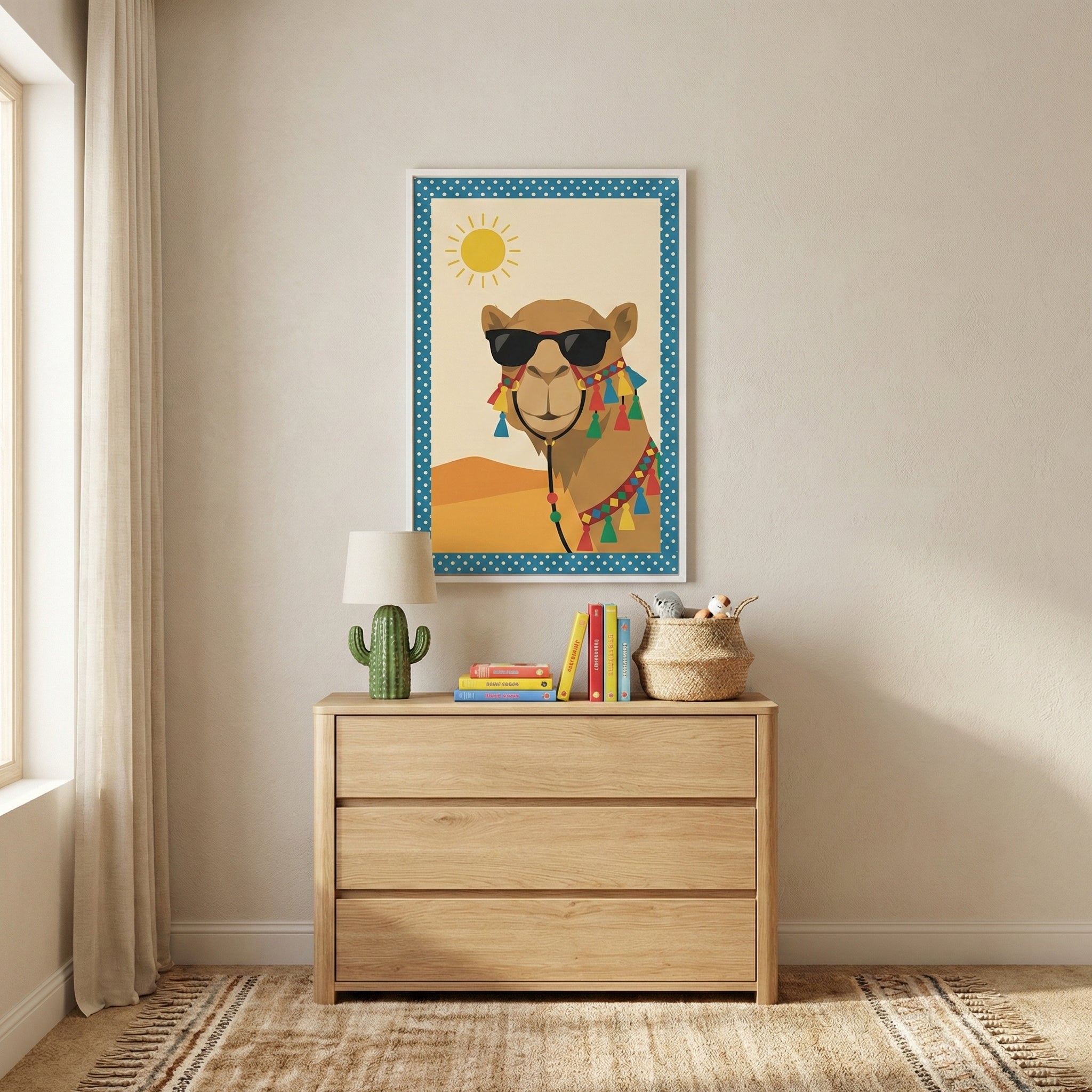 A minimalist children's room features a light wood three-drawer chest against a neutral off-white wall. On top of the chest sits a green cactus-shaped lamp with a white shade, a stack of colorful books, and a small woven basket. Centered on the wall above the chest is a framed illustration of a camel wearing dark sunglasses and colorful decorative tassels, set against a desert landscape with a bright sun and a blue polka-dot border. To the left, a long white curtain hangs beside a window, and a textured bei