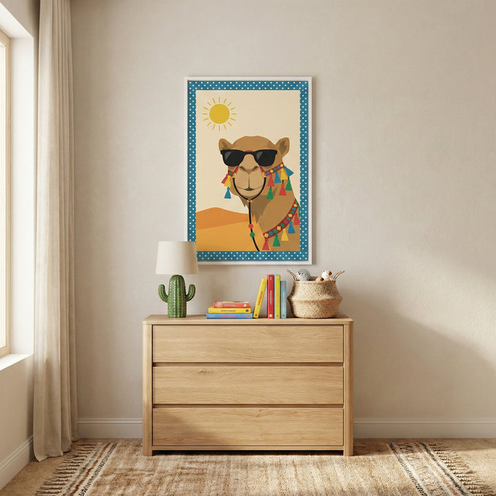 A minimalist children's room features a light wood three-drawer chest against a neutral off-white wall. On top of the chest sits a green cactus-shaped lamp with a white shade, a stack of colorful books, and a small woven basket. Centered on the wall above the chest is a framed illustration of a camel wearing dark sunglasses and colorful decorative tassels, set against a desert landscape with a bright sun and a blue polka-dot border. To the left, a long white curtain hangs beside a window, and a textured bei