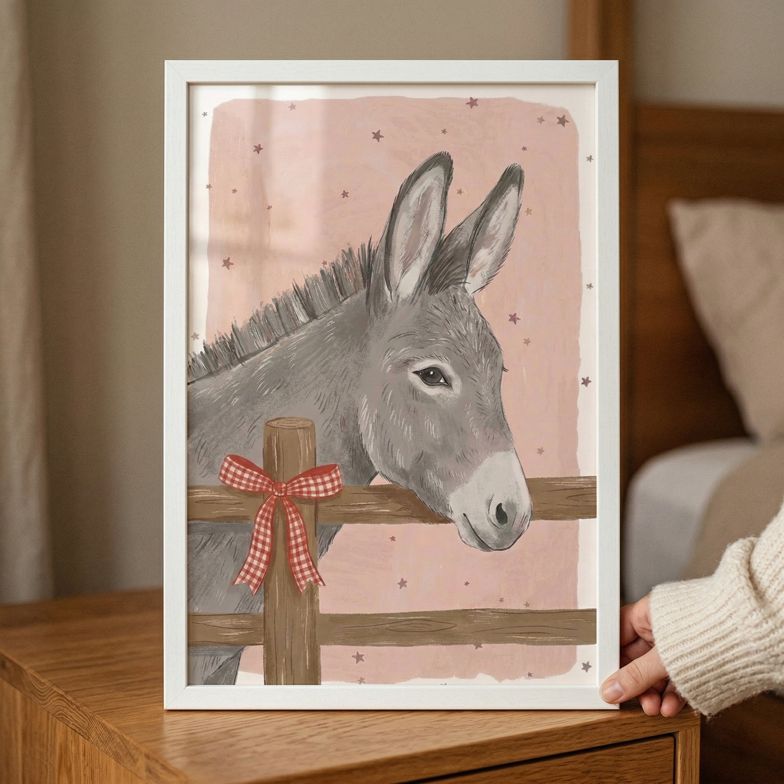 A framed illustration depicts a charming grey donkey looking over a wooden fence. The donkey has long ears and a soft expression, with its head resting near a post tied with a red and white checkered bow. The background is a delicate pink color, scattered with small brown stars, creating a gentle and whimsical scene. The artwork shows visible textured strokes, giving it a hand-crafted aesthetic. 