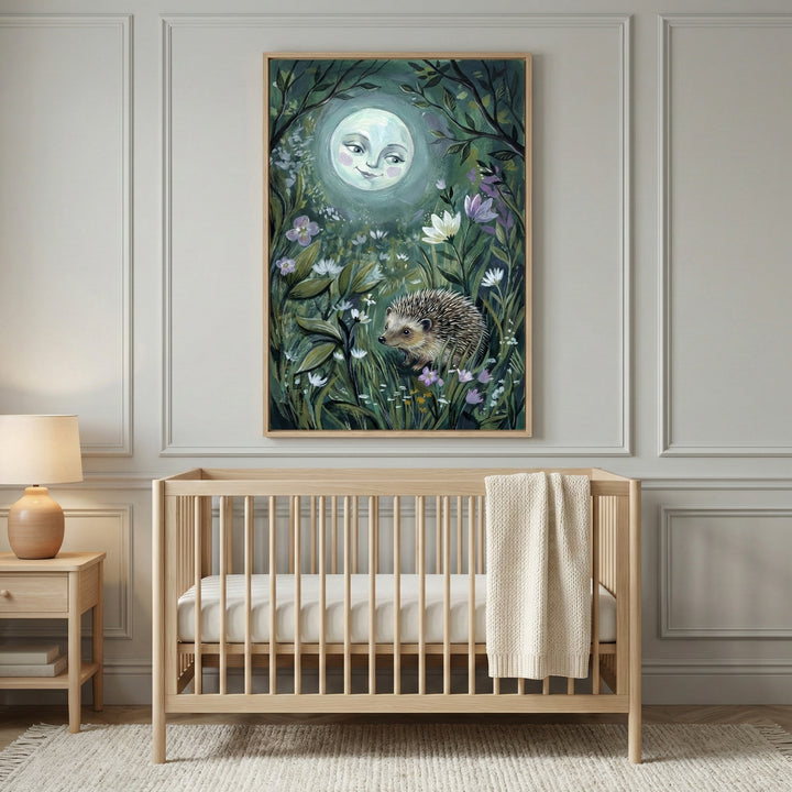 A minimalist nursery features a light wood crib and a large framed illustration centered on the wall. The illustration shows a whimsical scene with a personified moon smiling down at a hedgehog in a lush garden filled with flowers and foliage. A small wooden nightstand with a lamp stands to the left of the crib against light gray paneled walls.