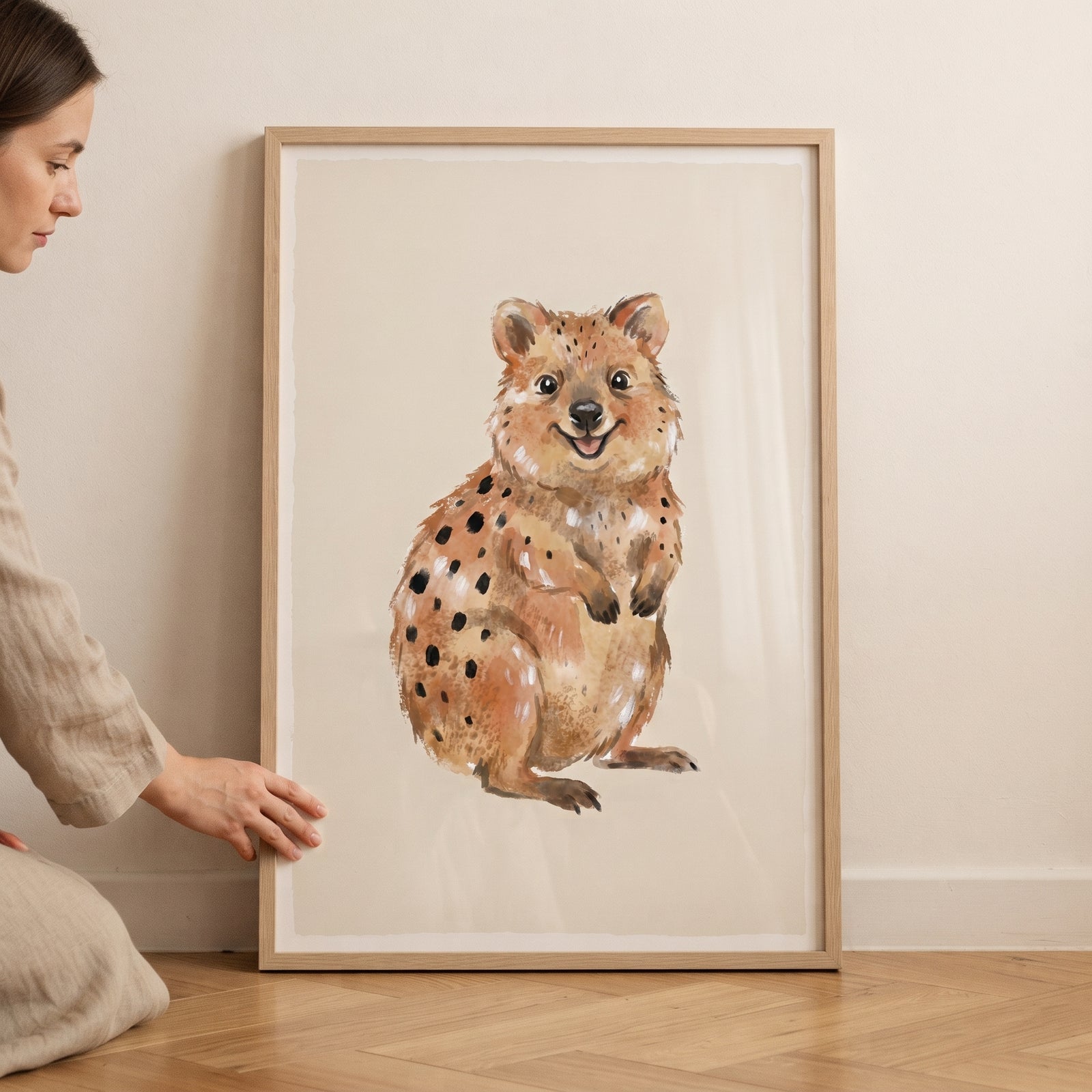 Woman holds a framed smiling watercolor brown quokka wall art against a light wall and wooden floor.