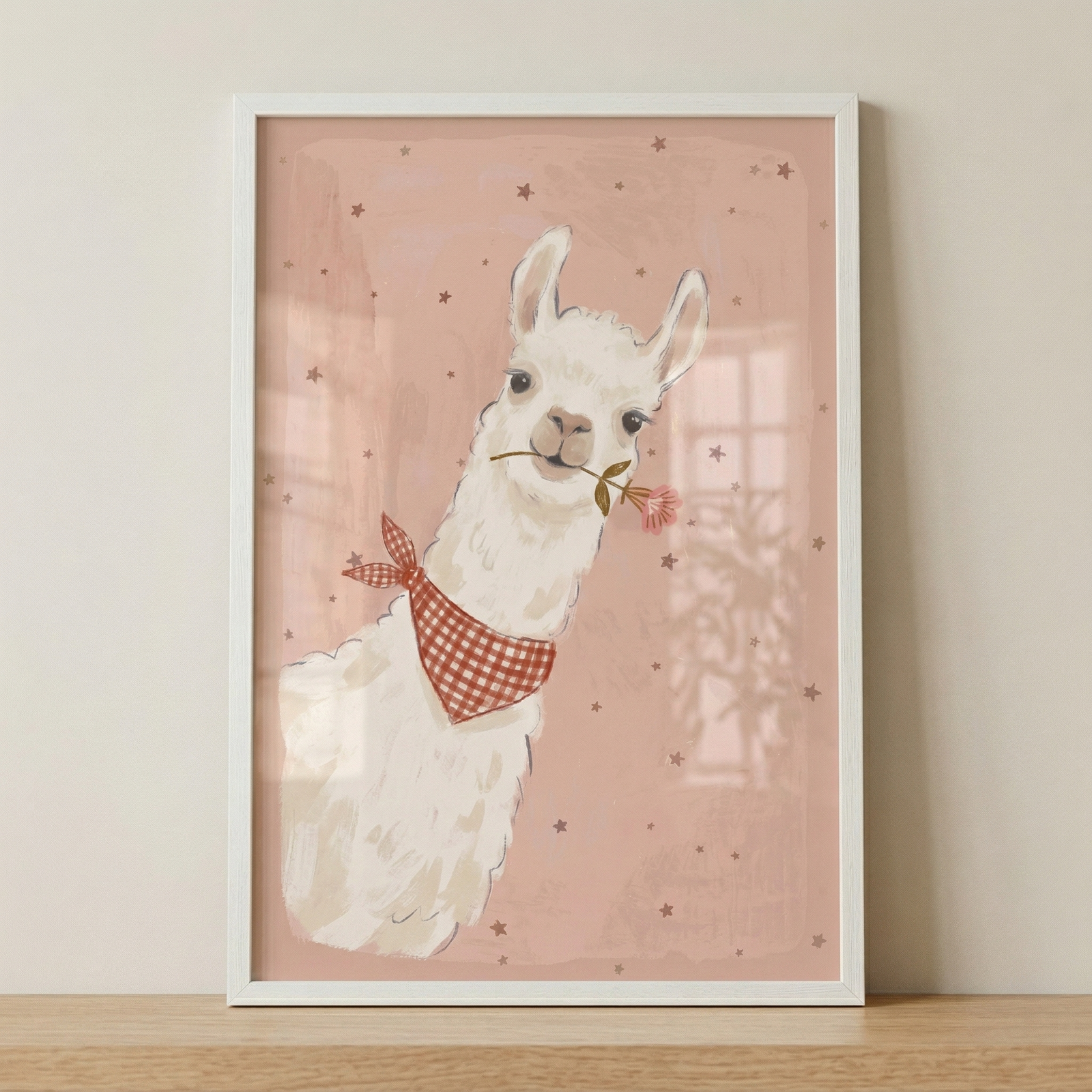 A framed vertical illustration depicts a friendly, white llama or alpaca against a soft pink background. The animal is adorned with a red and white checkered bandana around its neck and holds a small pink flower in its mouth. The background is sparsely decorated with tiny dark star shapes. The artwork is presented in a simple white frame, which rests on a light-toned wooden ledge against a plain, neutral wall. The style is whimsical and gentle, with a soft, hand-illustrated quality.