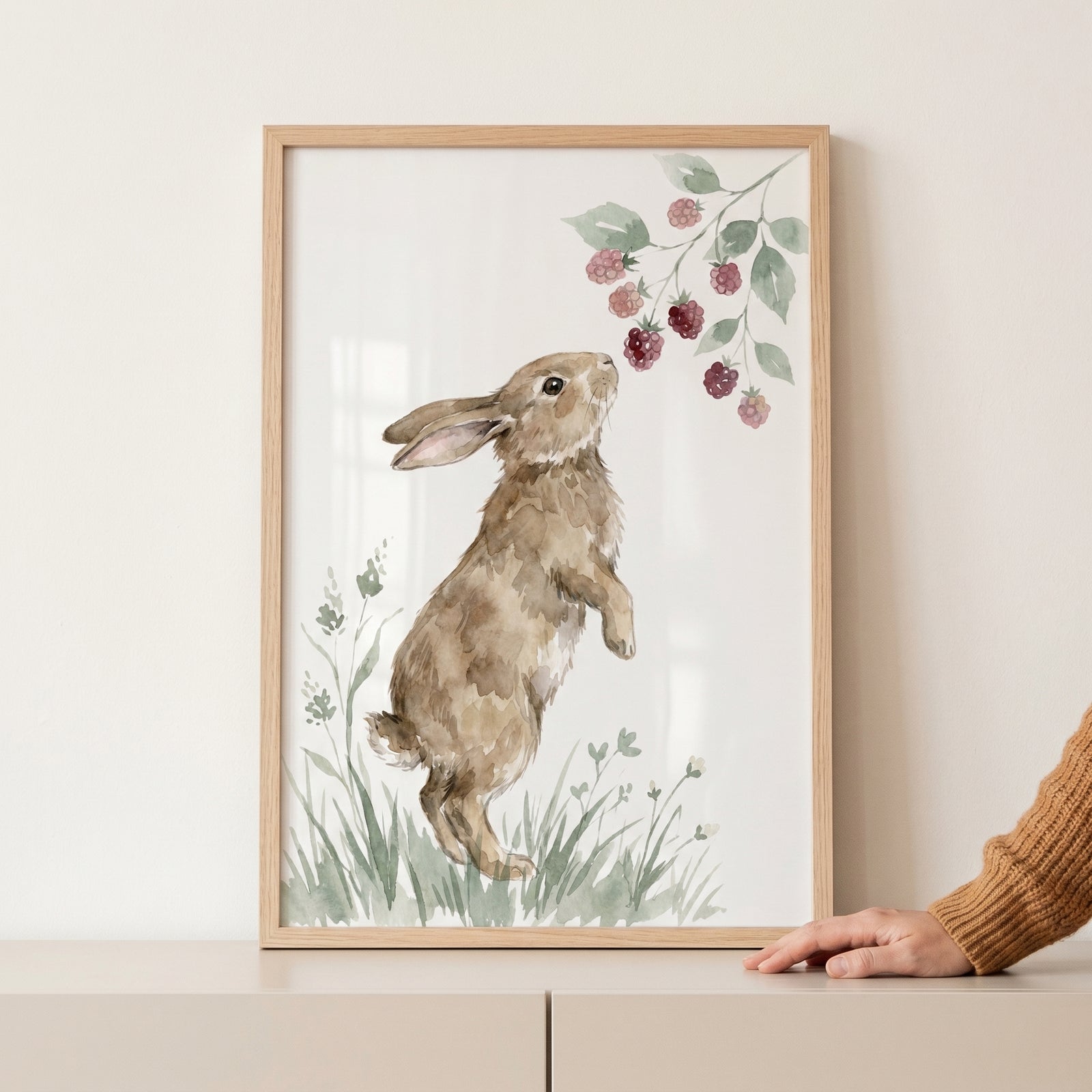 Close up of a framed watercolor rabbit standing on its hind legs reaching toward a branch of pink berries, with a hand resting beside the frame on a light surface.