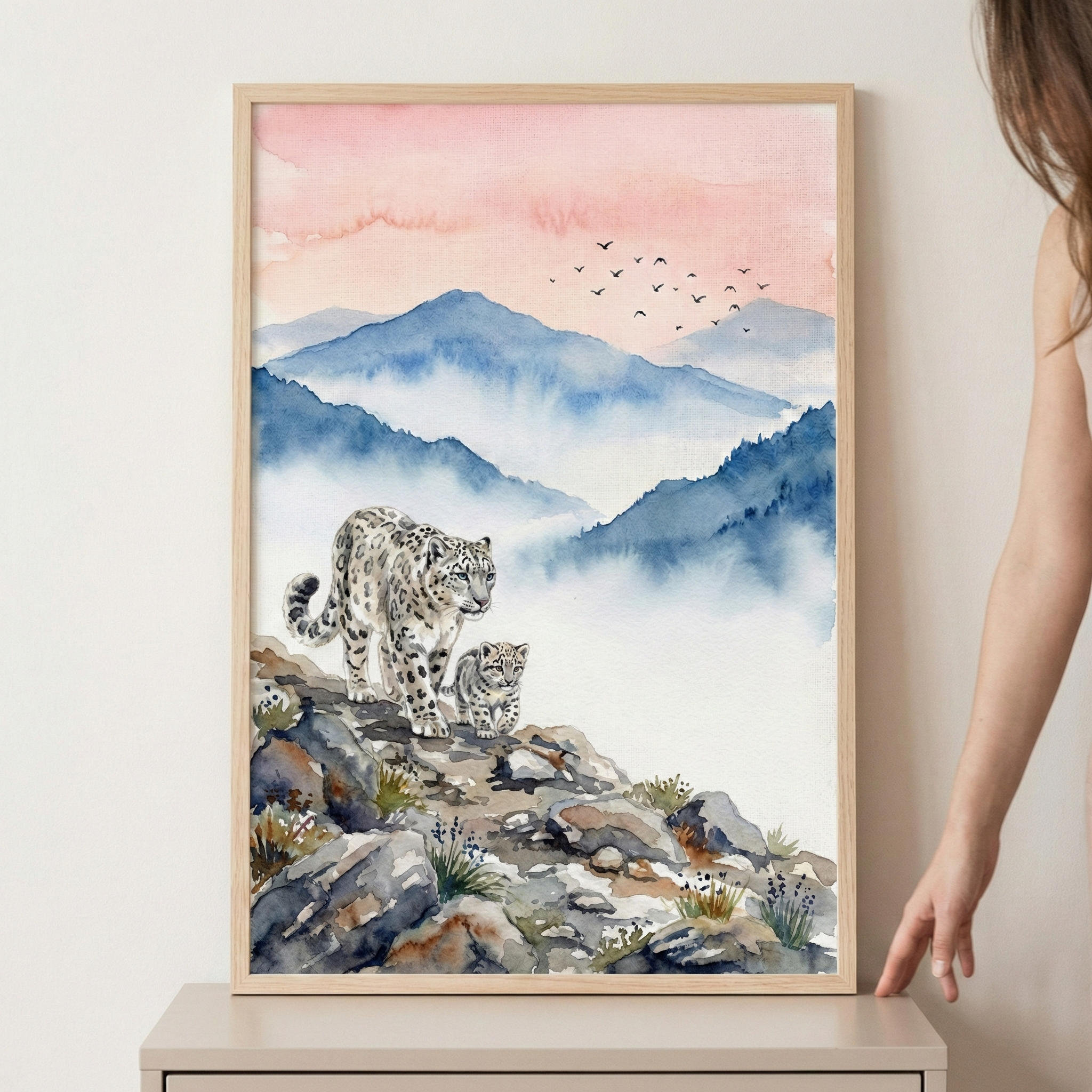A vertical, framed watercolor-style artwork features an adult snow leopard and its cub walking across a rocky mountain ridge. In the background, layers of blue-toned mountains are partially obscured by white mist, leading up to a soft peach and pink sky where a flock of small birds flies in the distance. The piece is encased in a light wood frame and sits on a neutral-toned surface. To the right, a person's hand rests near the base of the frame, with their long hair and shoulder visible at the edge.